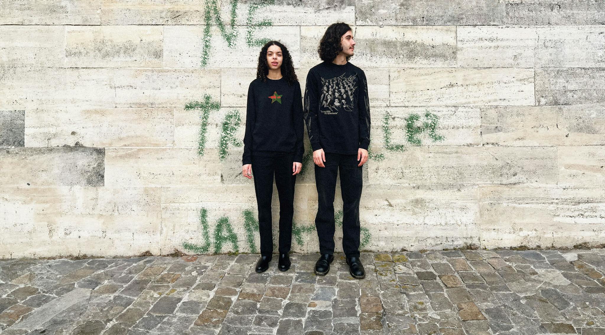 Two individuals in black attire against a graffitied wall.