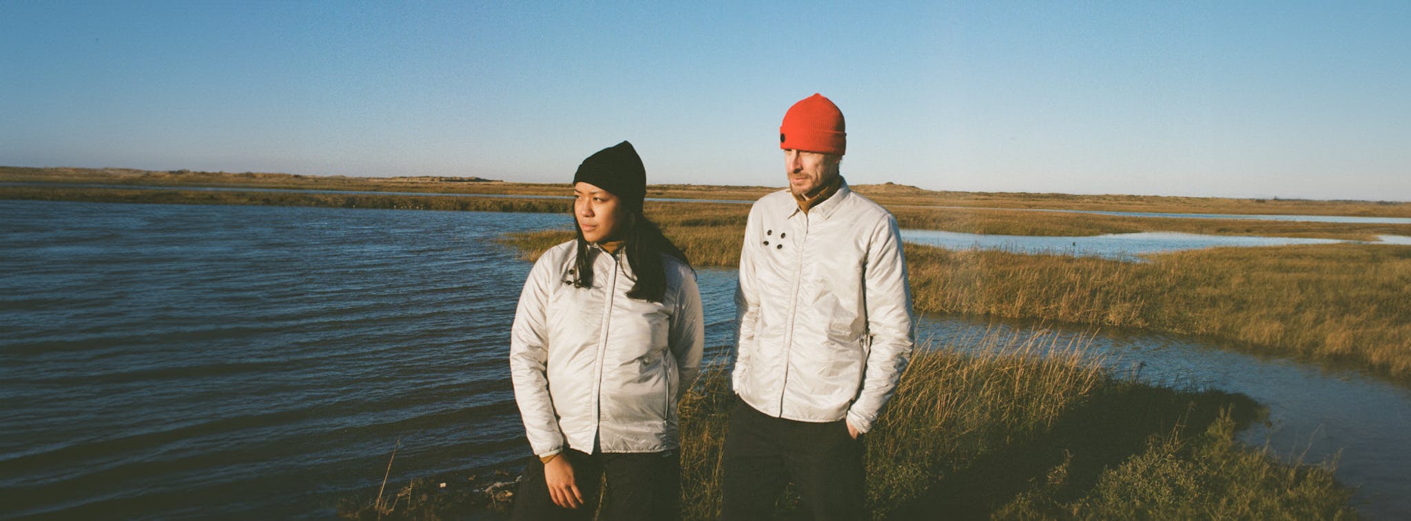 Two individuals in silver jackets and beanies, one black and one red, standing in the late afternoon sun by a marshland.