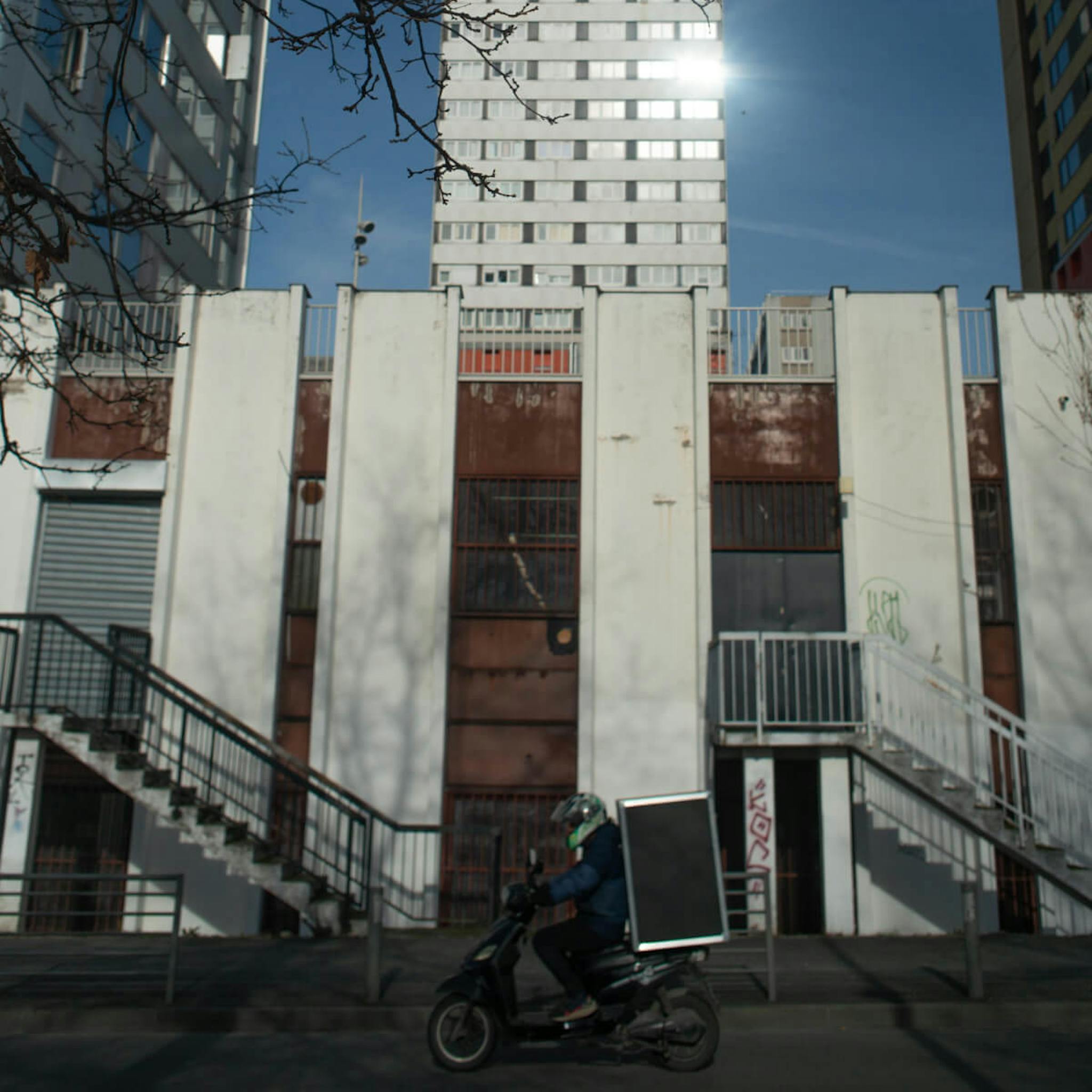 A delivery person on a scooter carrying a large insulated box passes in front of a stark urban building