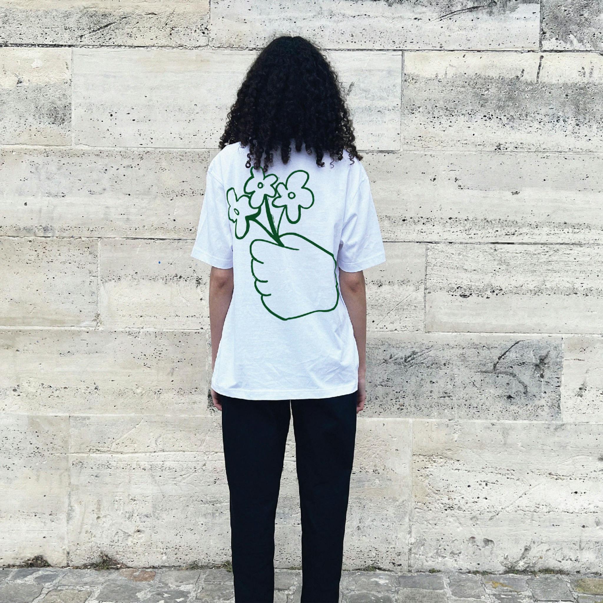 A woman with long dark hair wearing a white T-shirt with a green outline drawing of a hand holding three flowers on the back.