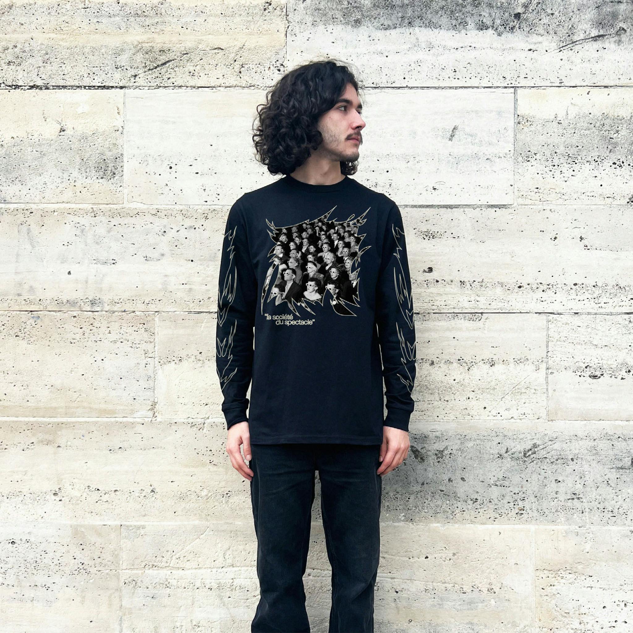 A man with long dark hair wearing a black long-sleeve T-shirt with a graphic of an audience with glasses and the text "la société du spectacle."