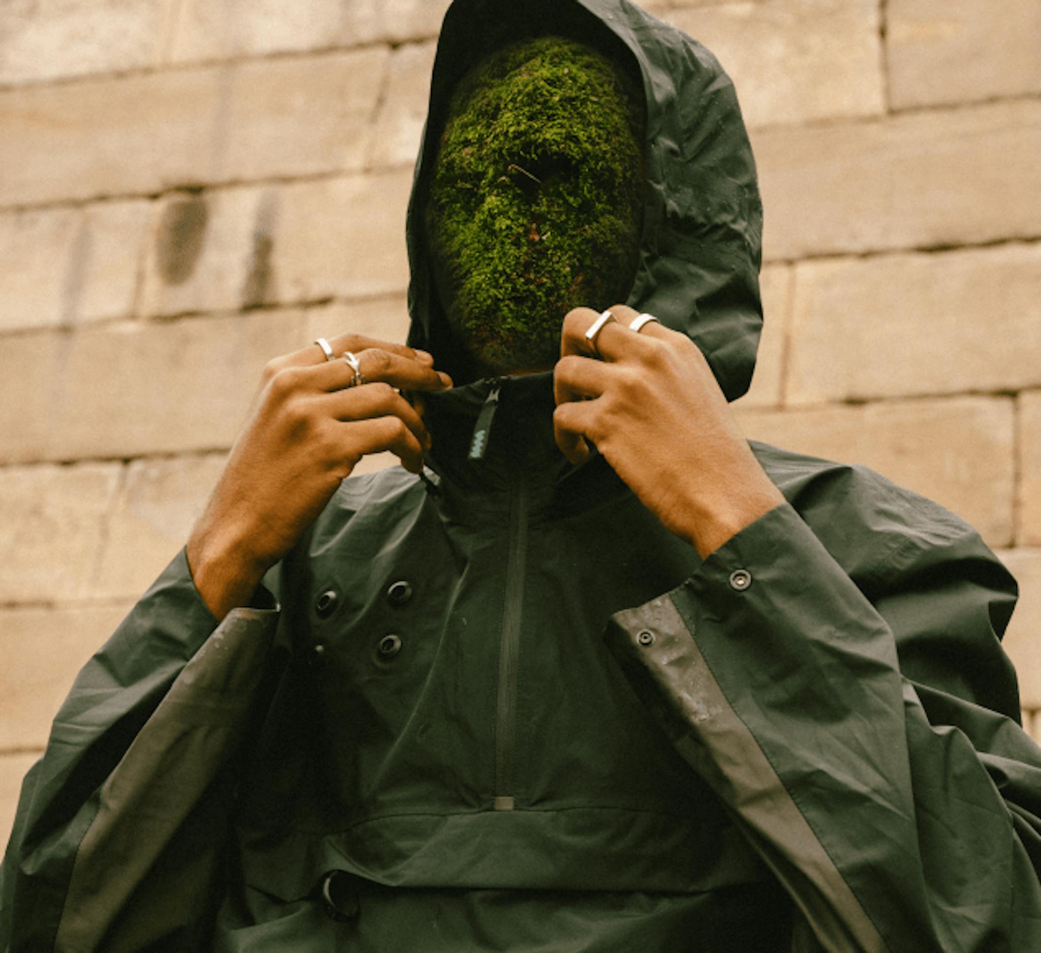 A person in a dark green hooded jacket with a moss-covered head instead of a human face, blending elements of nature with human form.