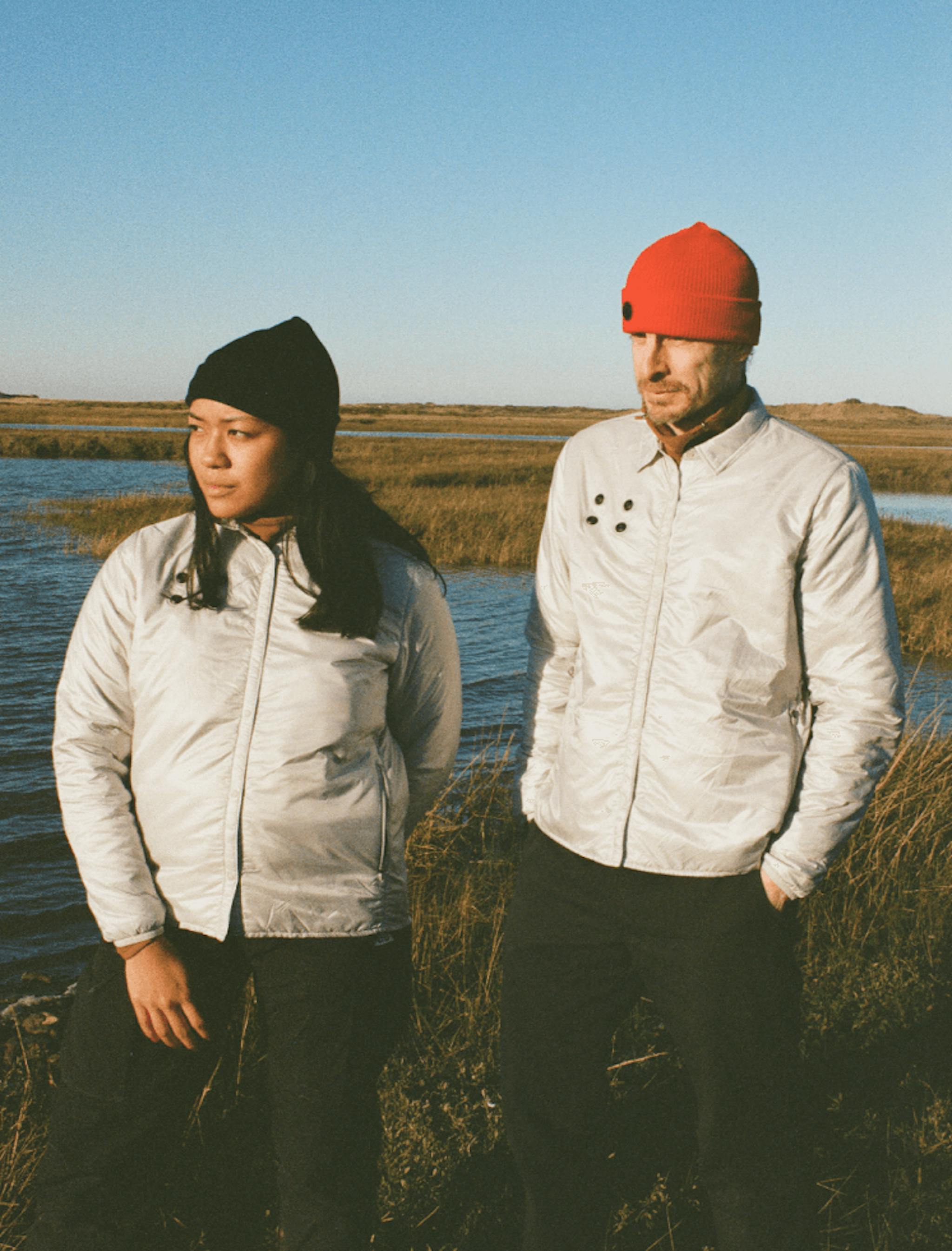 Two individuals in silver jackets and beanies, one black and one red, standing in the late afternoon sun by a marshland.