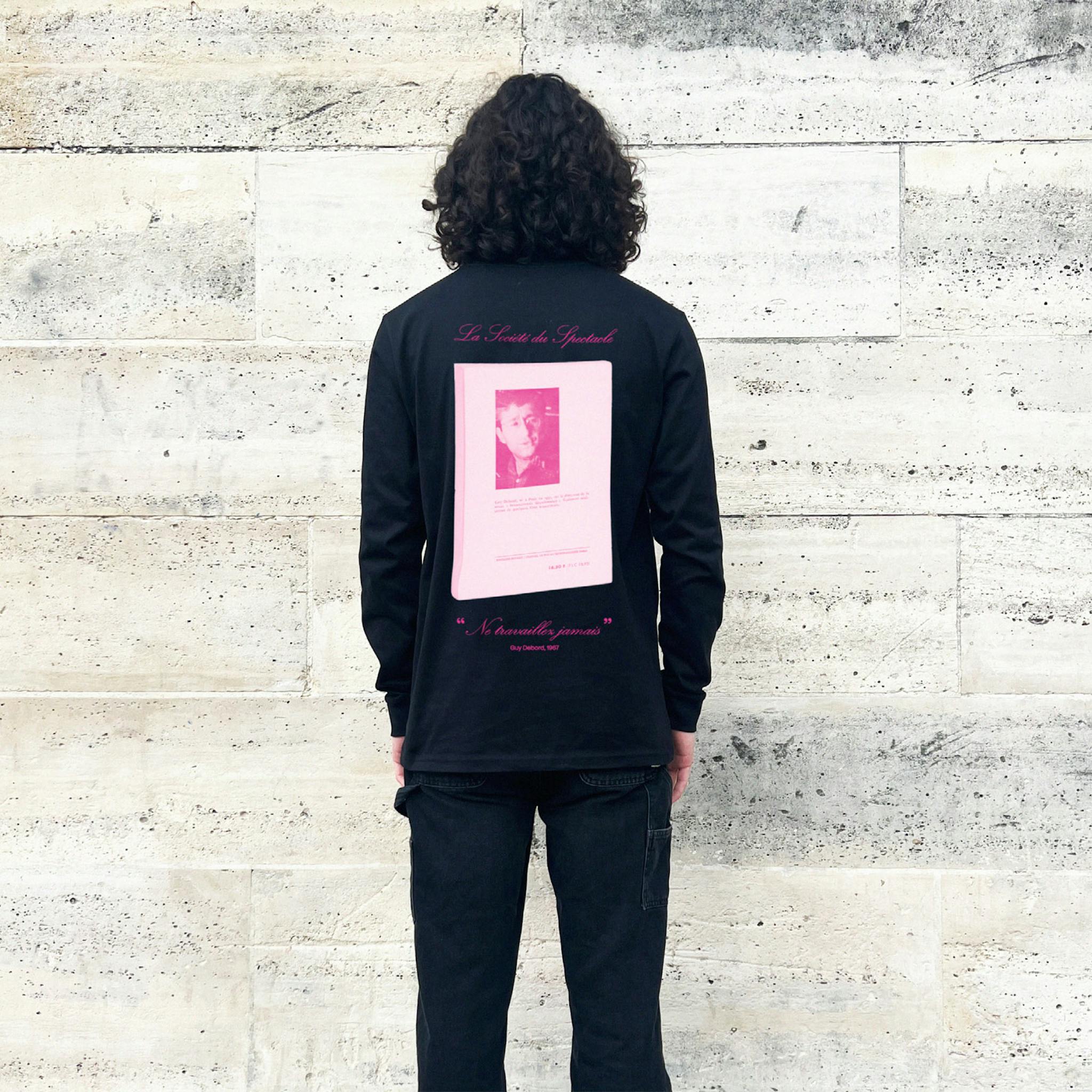 A man with long dark hair wearing a black long-sleeve T-shirt with large pink book graphic on the back and french words over and underneath.