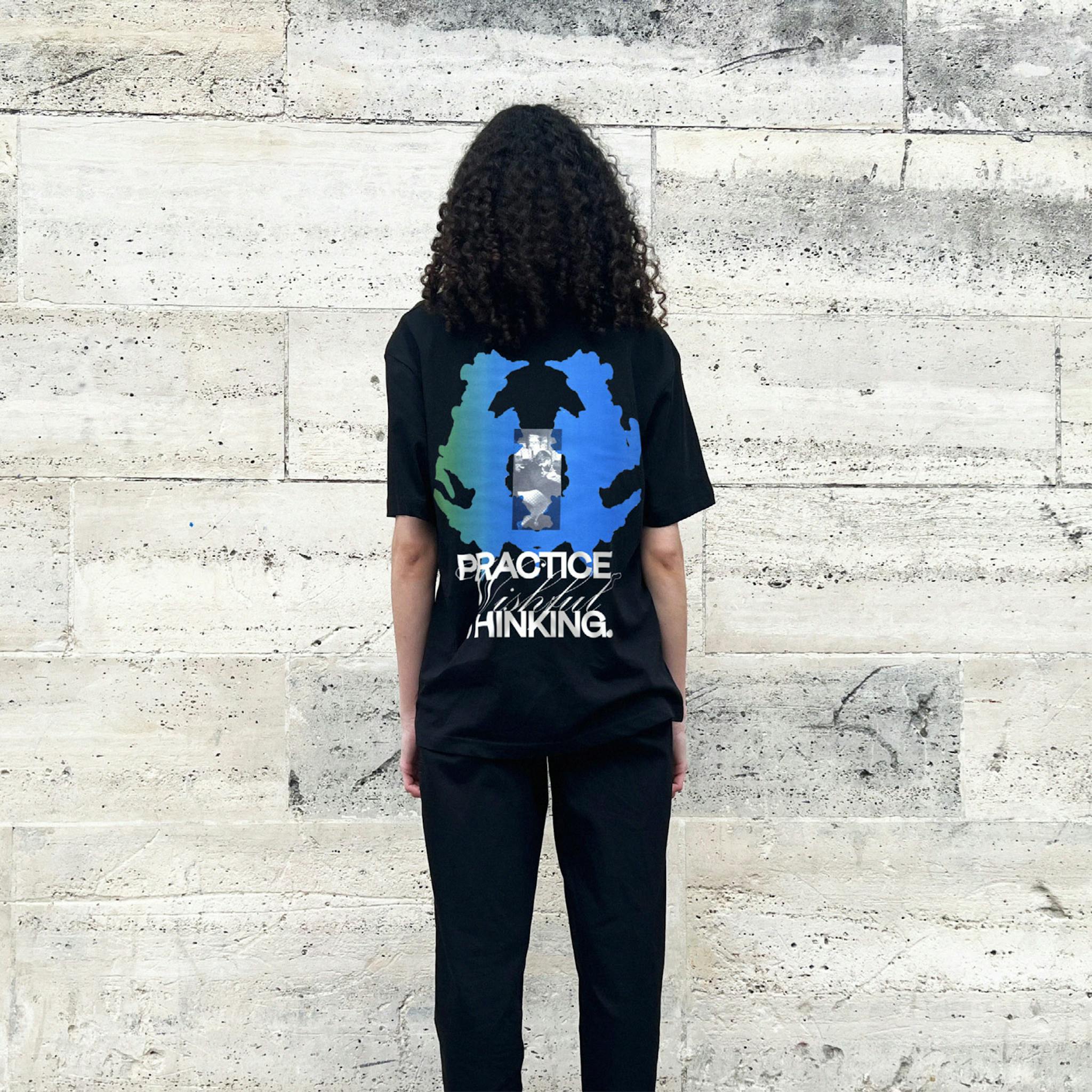 A woman with long dark hair wearing black T-shirt with a large blue and green graphic and text on the back saying "Practice Wishful Thinking"