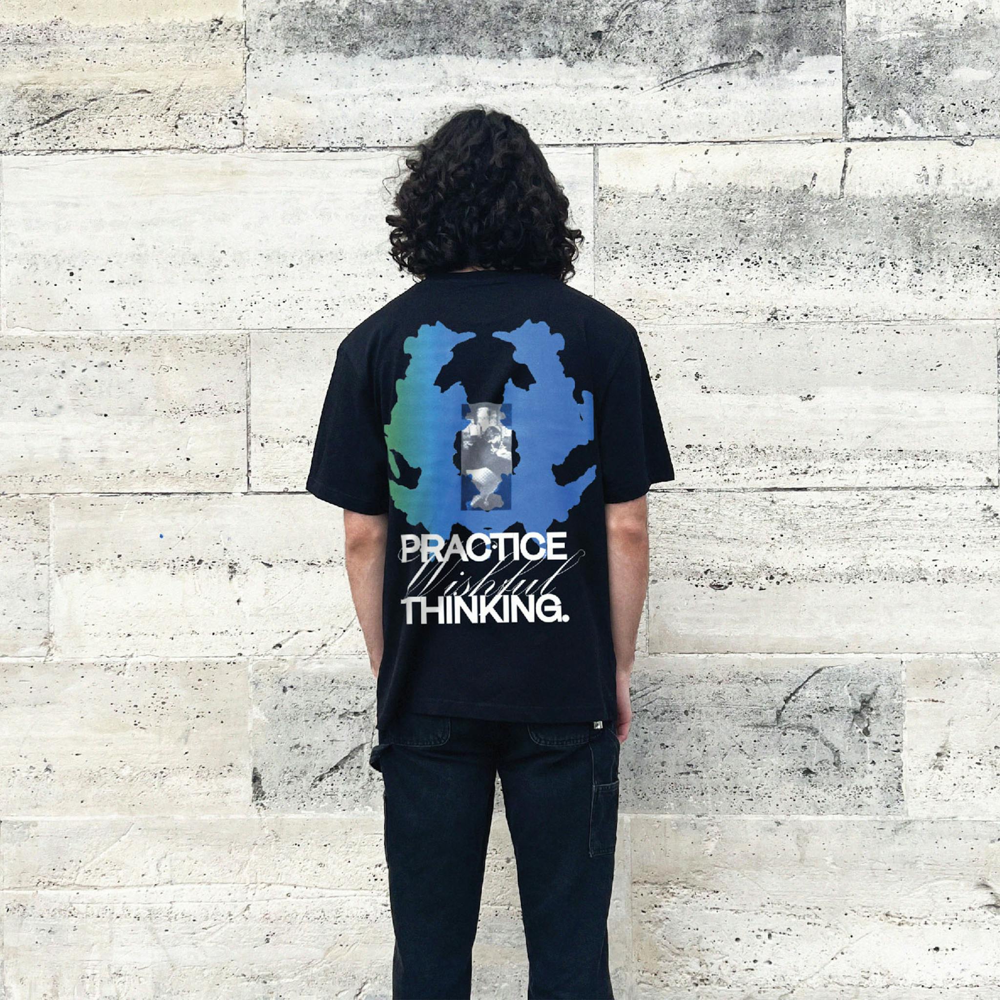A man with long dark hair wearing black T-shirt with a large blue and green graphic and text on the back saying "Practice Wishful Thinking"