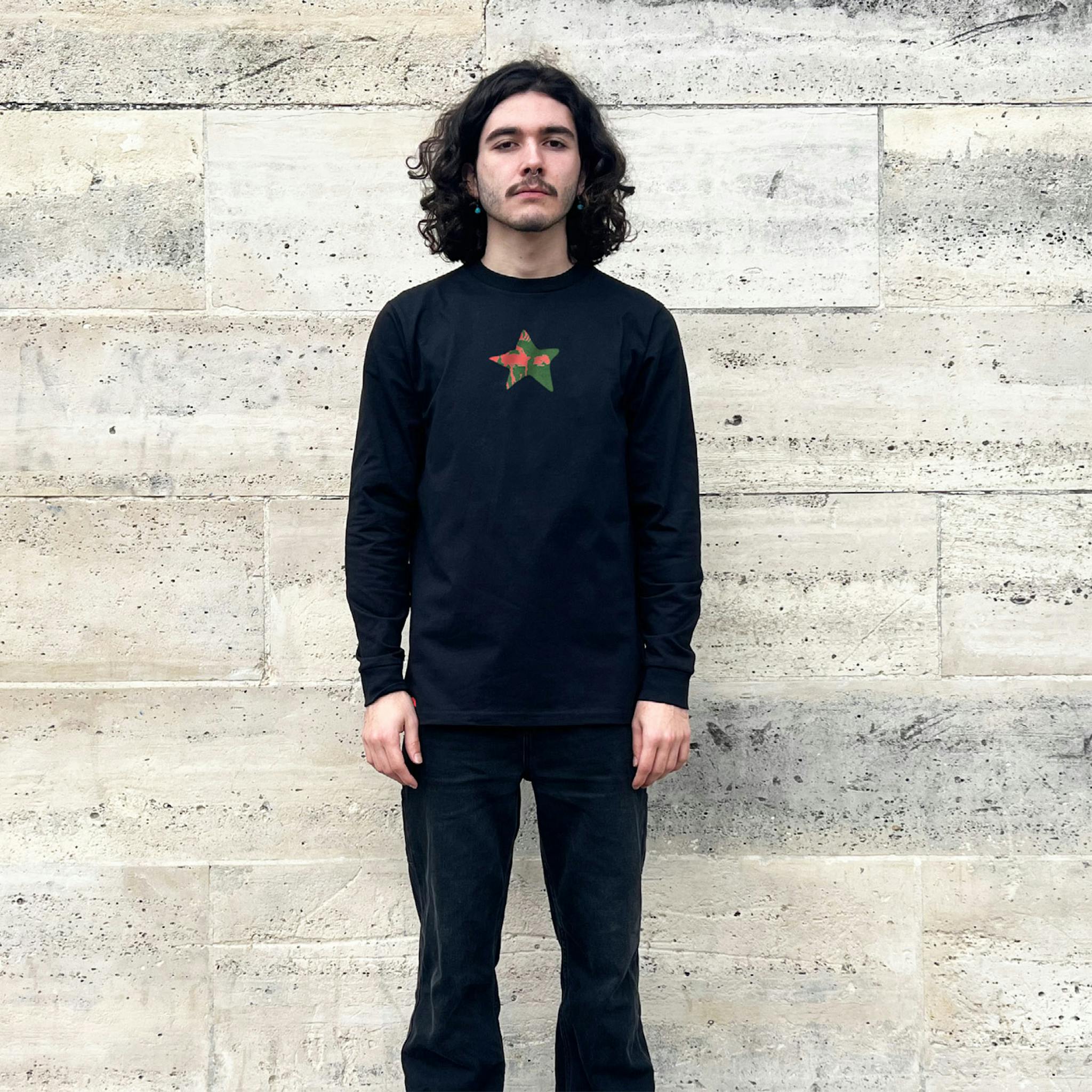 A man with long dark hair wearing a black long-sleeve T-shirt with a small graphic of a red and green star on the chest.