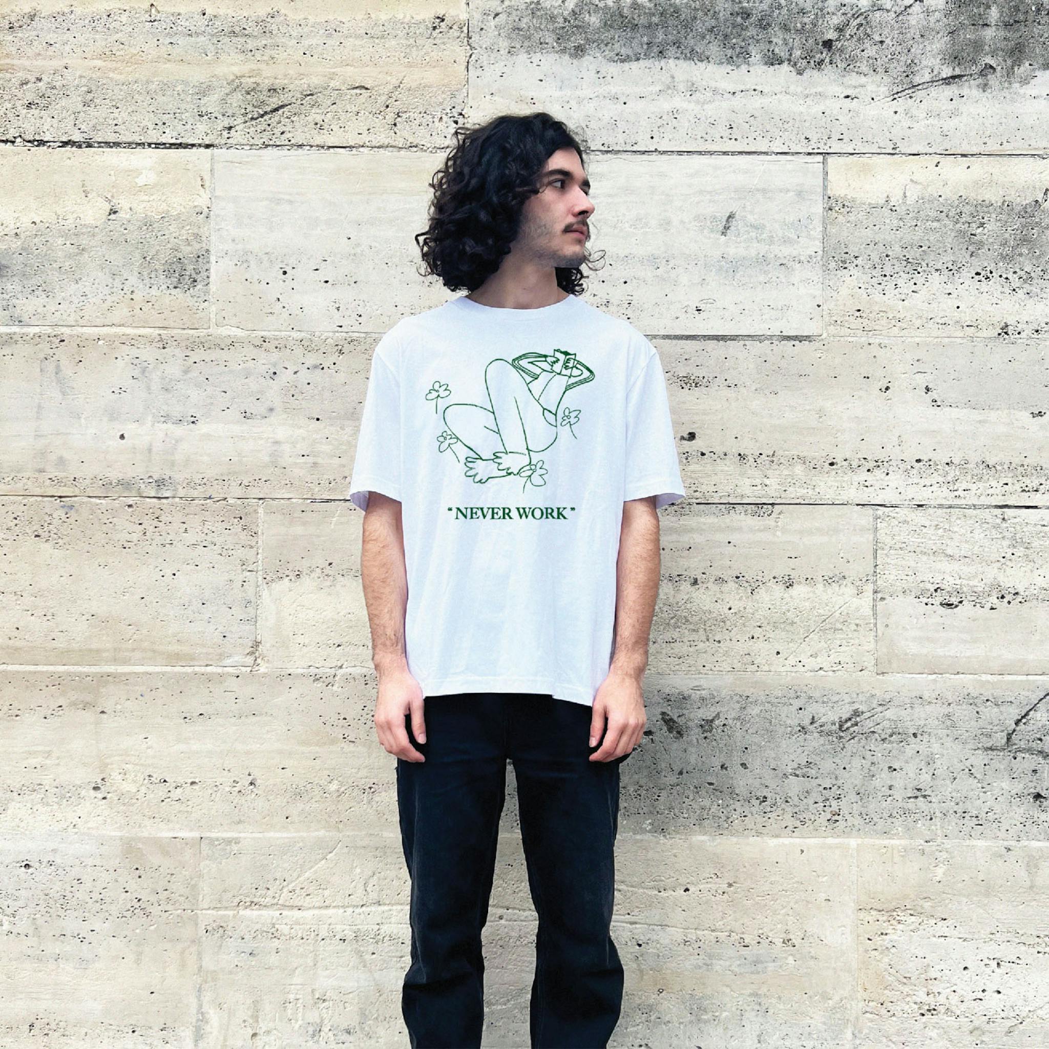 A man with long dark hair wearing a white T-shirt with a green outline drawing of a figure lying back with legs up, surrounded by flowers, and the phrase "NEVER WORK" below it.