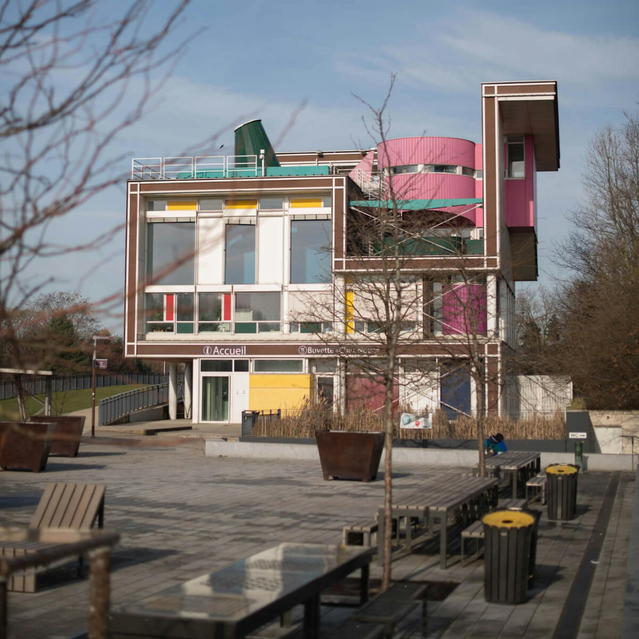 A contemporary building with pink and yellow panels, surrounded by bare trees and outdoor benches.
