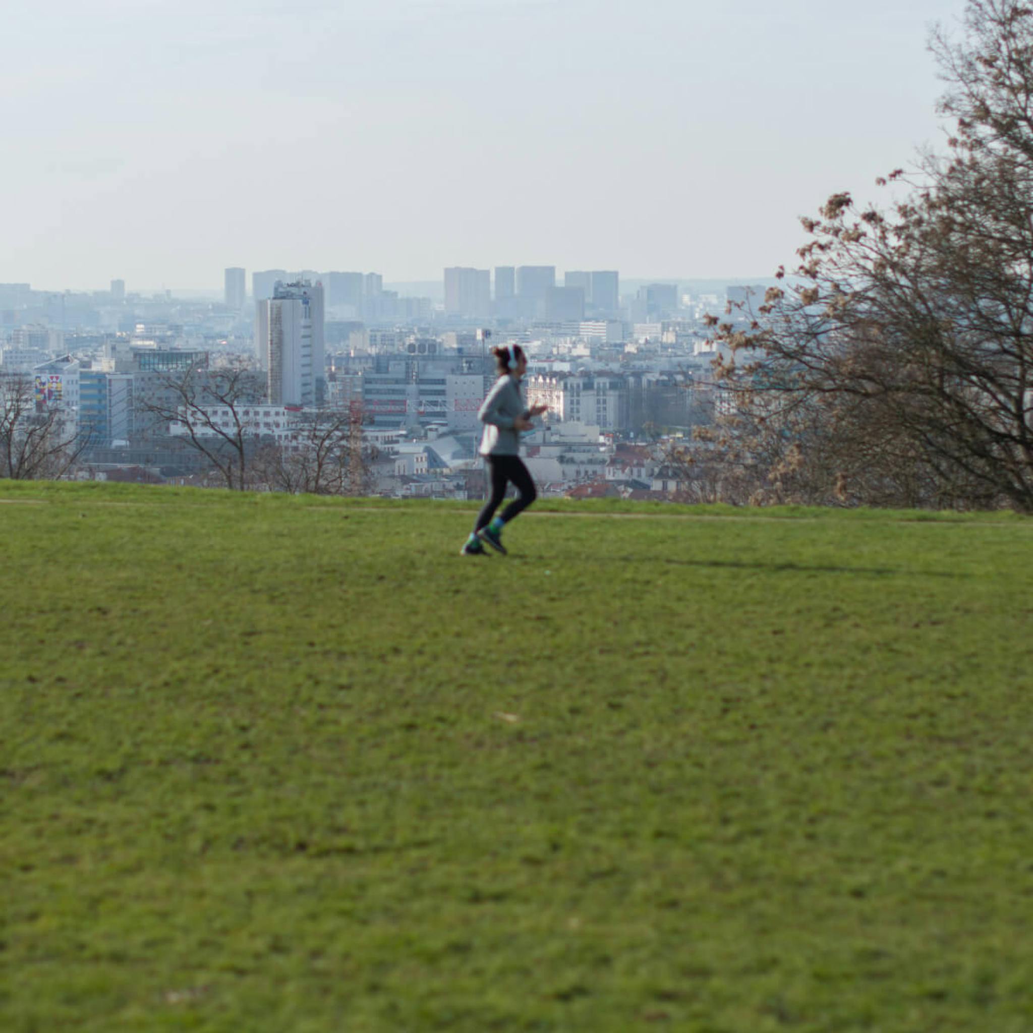 A jogger runs across a grassy hill with the sprawling cityscape of Paris in the background.