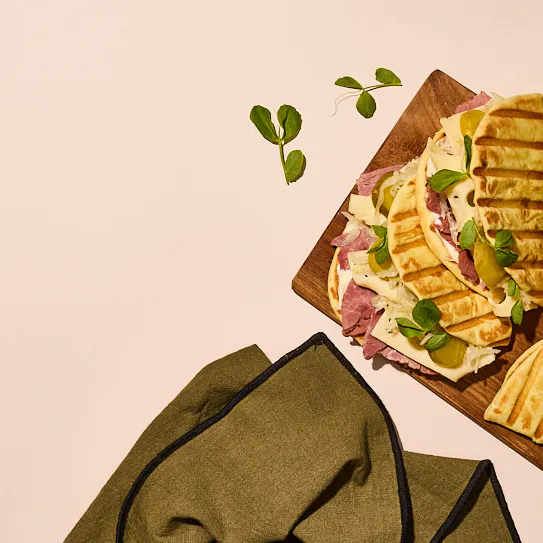 Overhead view of grilled flatbread pieces arranged on a wooden board and layered with slices of cured meat, shaved cheese, and small green leaves. A folded green napkin sits in the lower left on a neutral beige surface, with a few scattered leaves adding detail around the composition.