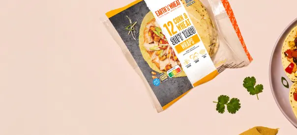 A packaged product of “Earth & Wheat 10 Corn & Wheat Soft Taco Wraps” lies on a beige surface. The packaging is partially visible, showing an image of filled tacos along with branding and product text. A few coriander leaves are scattered nearby, and the edge of a plate with tacos appears on the right side.
