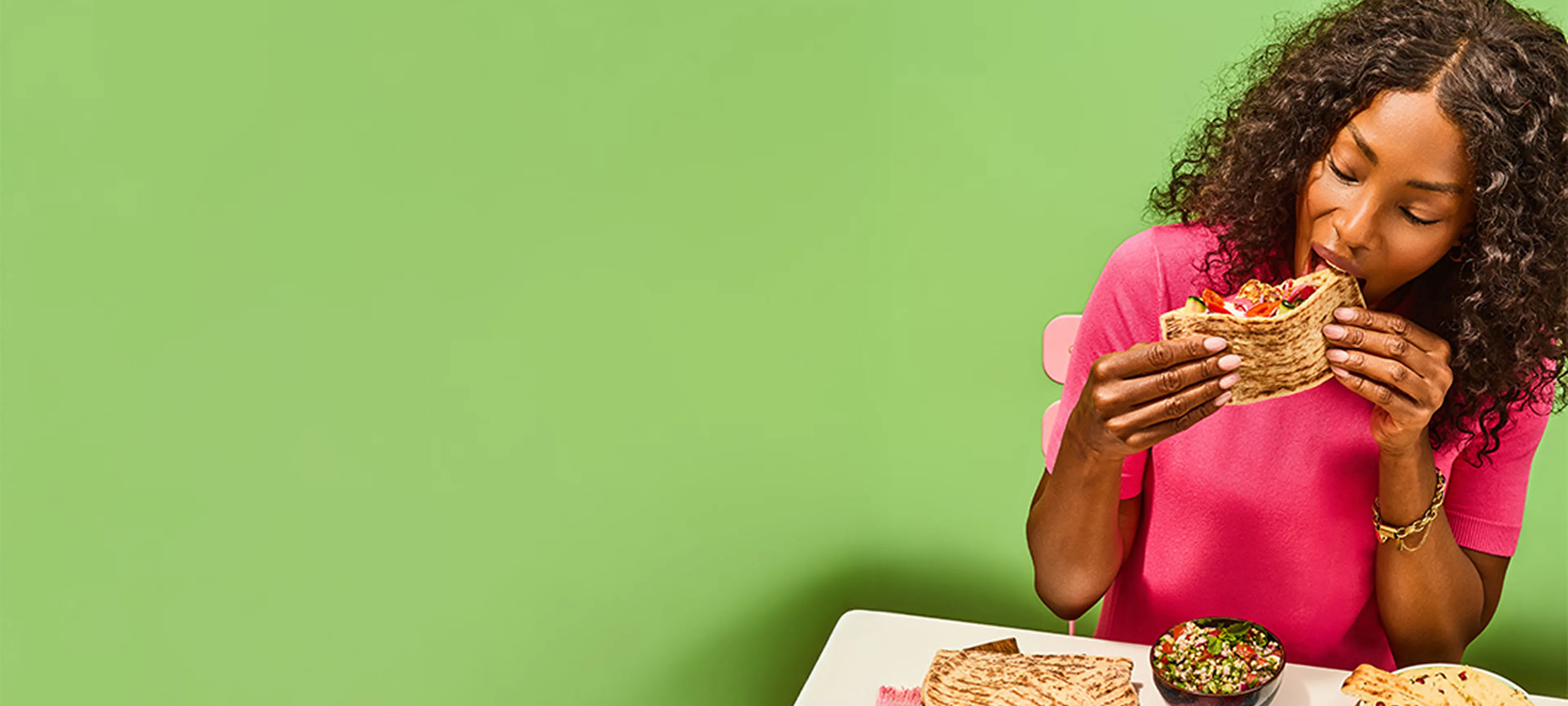 Person in a bright pink top holding and eating a stuffed flatbread sandwich filled with colourful vegetables, seated at a table against a solid green background. On the table are stacked flatbreads, a bowl of salsa, and other food items arranged for a vibrant meal scene.