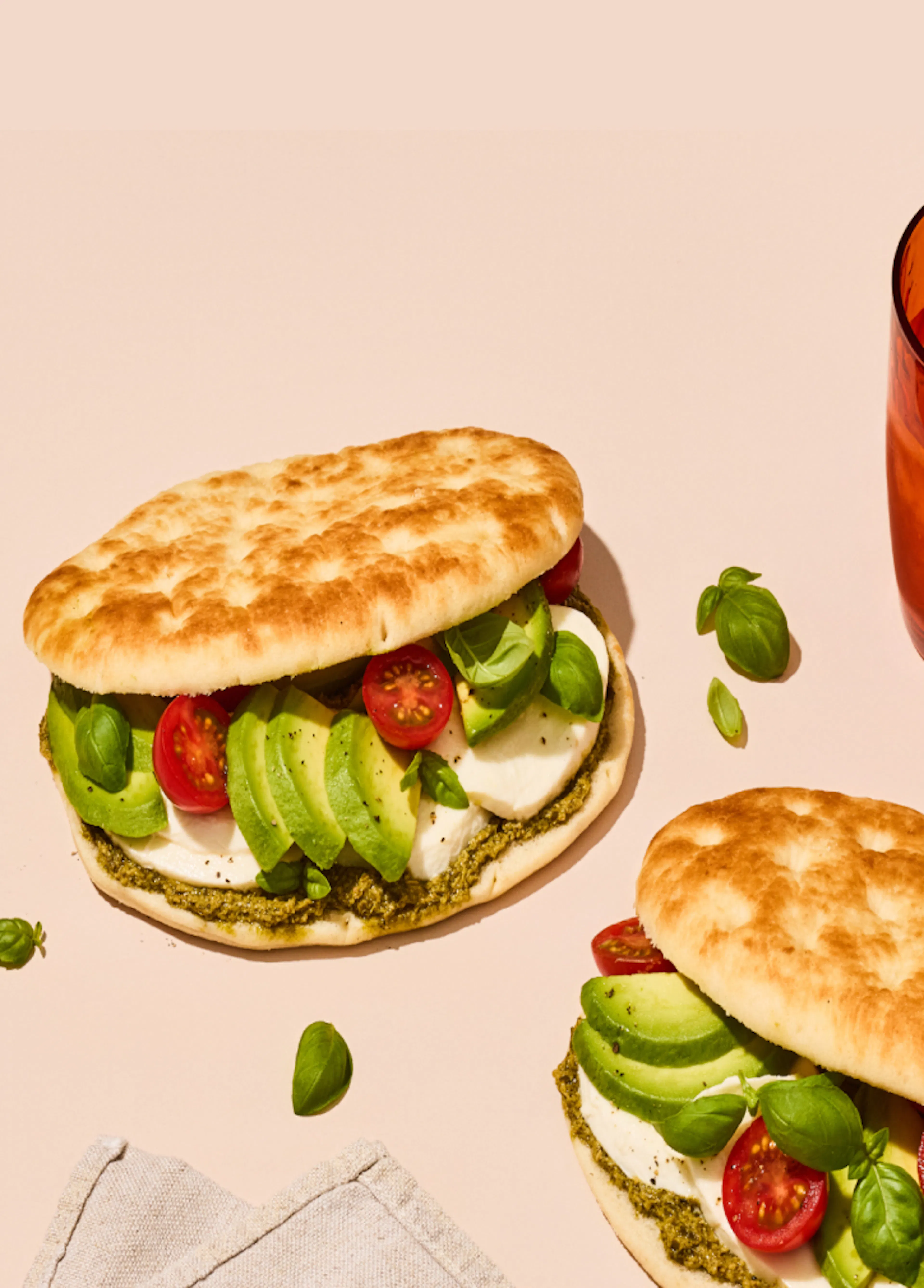 Two folded flatbread sandwiches filled with sliced avocado, cherry tomatoes, leafy greens, and soft white cheese sit on a beige surface. A spread of green pesto is visible along the bread. An amber glass with an iced orange drink is placed in the upper right, while a folded napkin rests in the lower left. Small basil leaves are scattered around the scene.