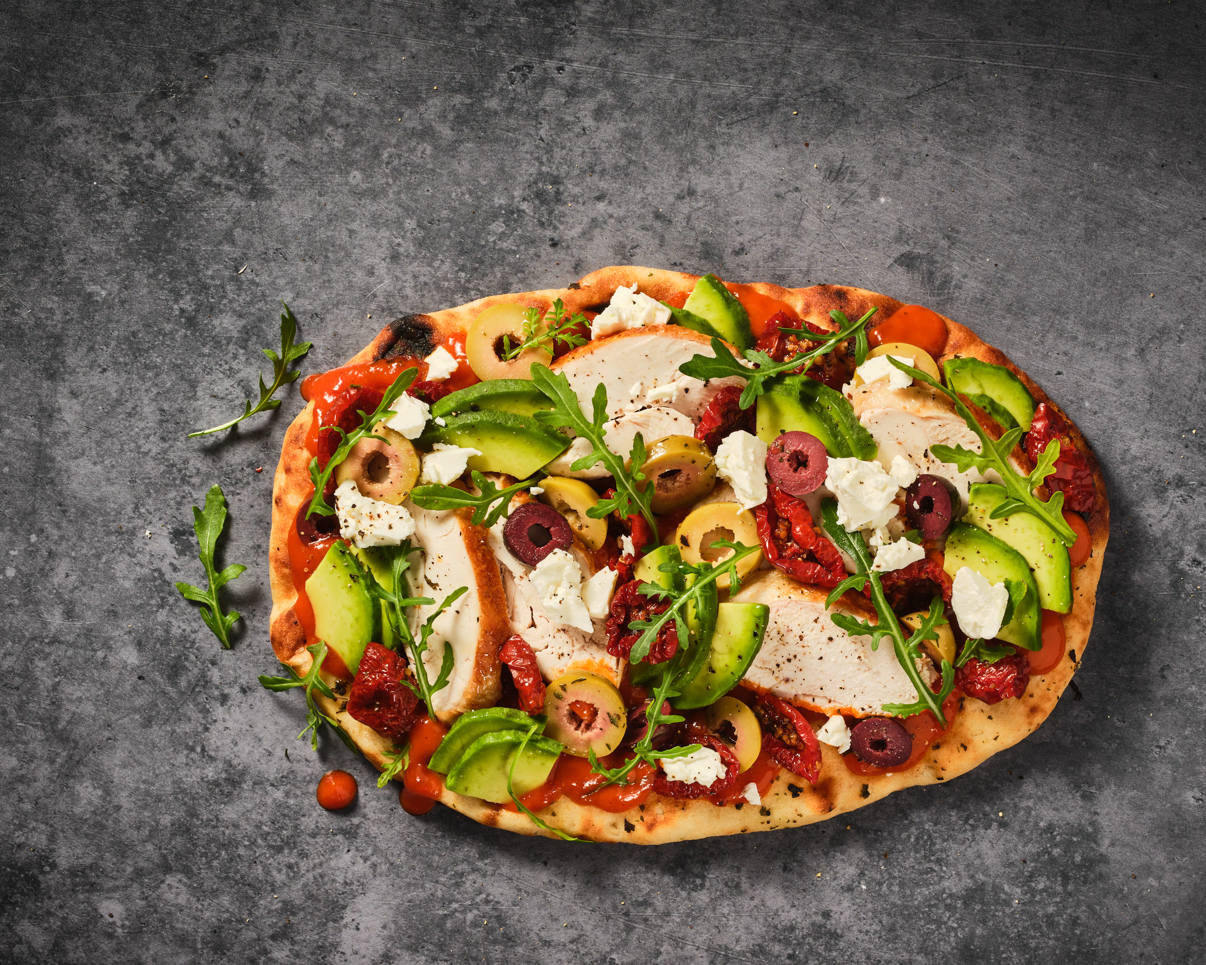 Overhead view of an naan bread topped with sliced avocado, olives, sun-dried tomatoes, soft white cheese, and fresh rocket leaves over a layer of tomato sauce. The flatbread sits on a dark grey textured surface with a few scattered herbs and small tomato pieces nearby.