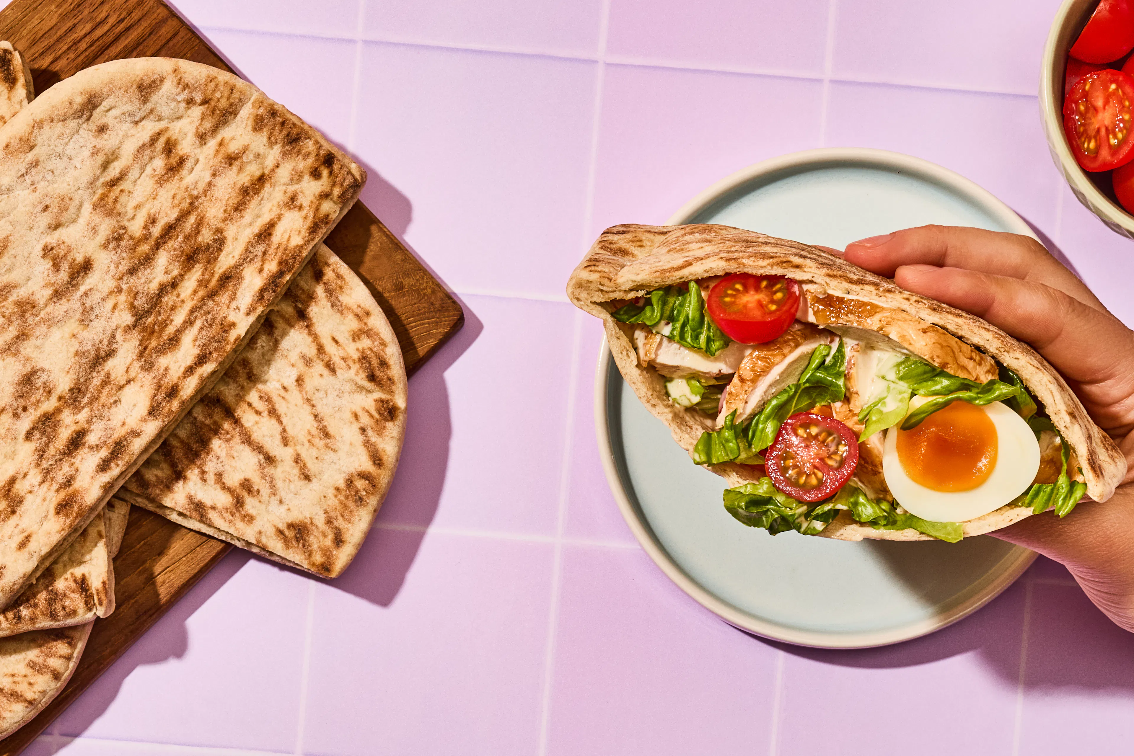 Chicken, egg filling in an open pitta pocket being held to the left on a lilac tiled background.