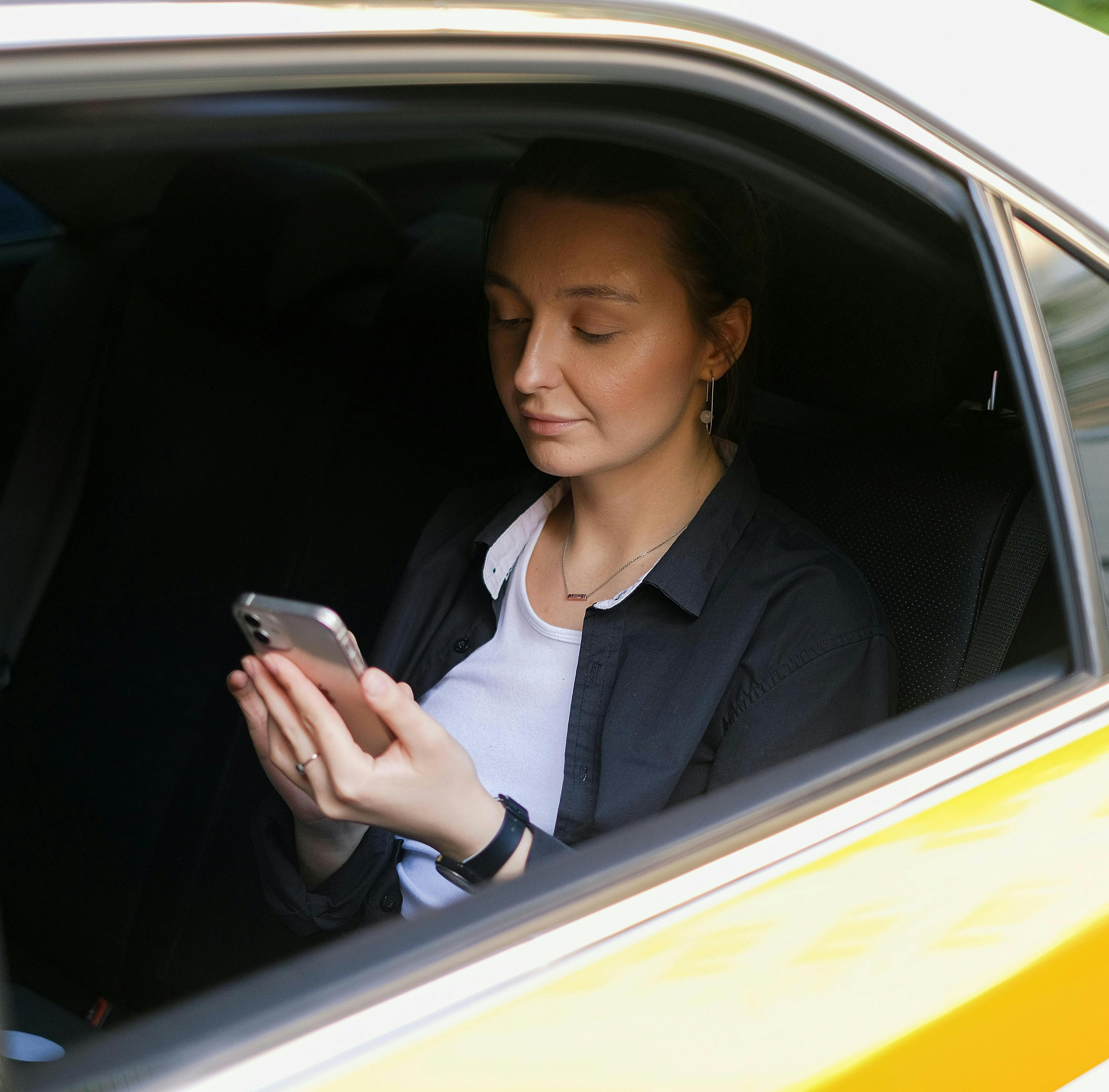 на зображенні жінка з телефоном в руках сидить на задньому сидінні жовтого авто