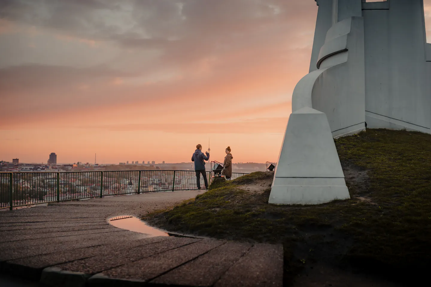 Two people at the railing below the white Three Crosses monument, looking out over Vilnius at sunset
