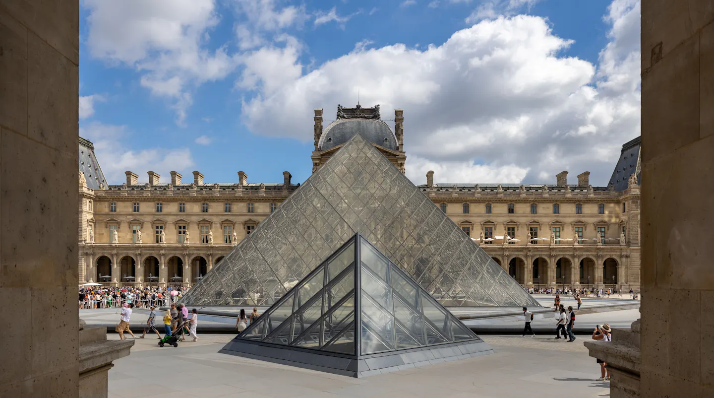 The glass pyramid and the wings of the Louvre framed between two stone columns of the palace arcade, under a blue sky with billowing white cloud