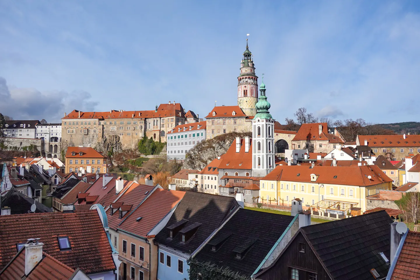 Cesky Krumlov's painted round castle tower and the long chateau range standing above a jumble of red-tiled old town roofs, with the white church tower beside them under a blue sky