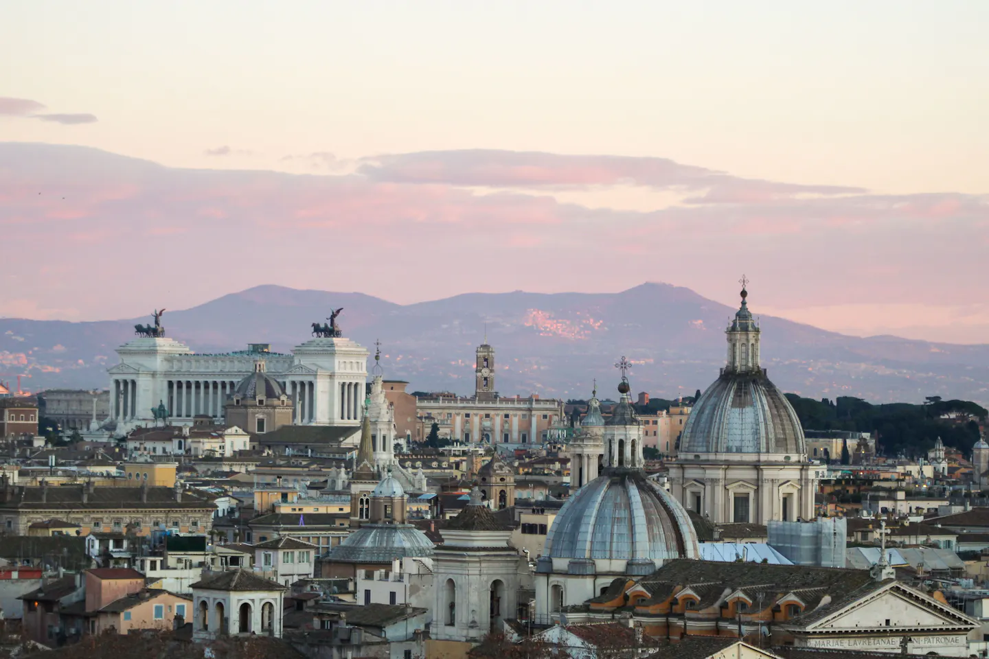 Rome's skyline at dusk under a pink and apricot sky: the domes of Sant'Andrea della Valle and Sant'Agnese, the white marble of the Vittoriano and the Alban hills layered behind the rooftops