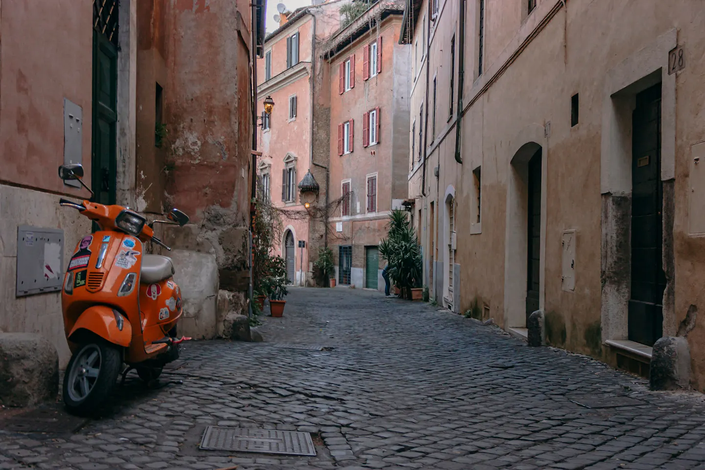 A quiet cobbled lane in central Rome between faded ochre and pink houses, with potted plants and an orange Vespa parked by the wall