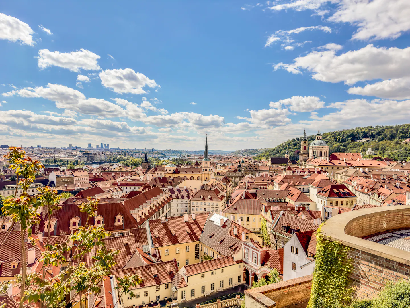 Terracotta rooftops and green church domes of Prague's Mala Strana below a wooded hill on a bright autumn day