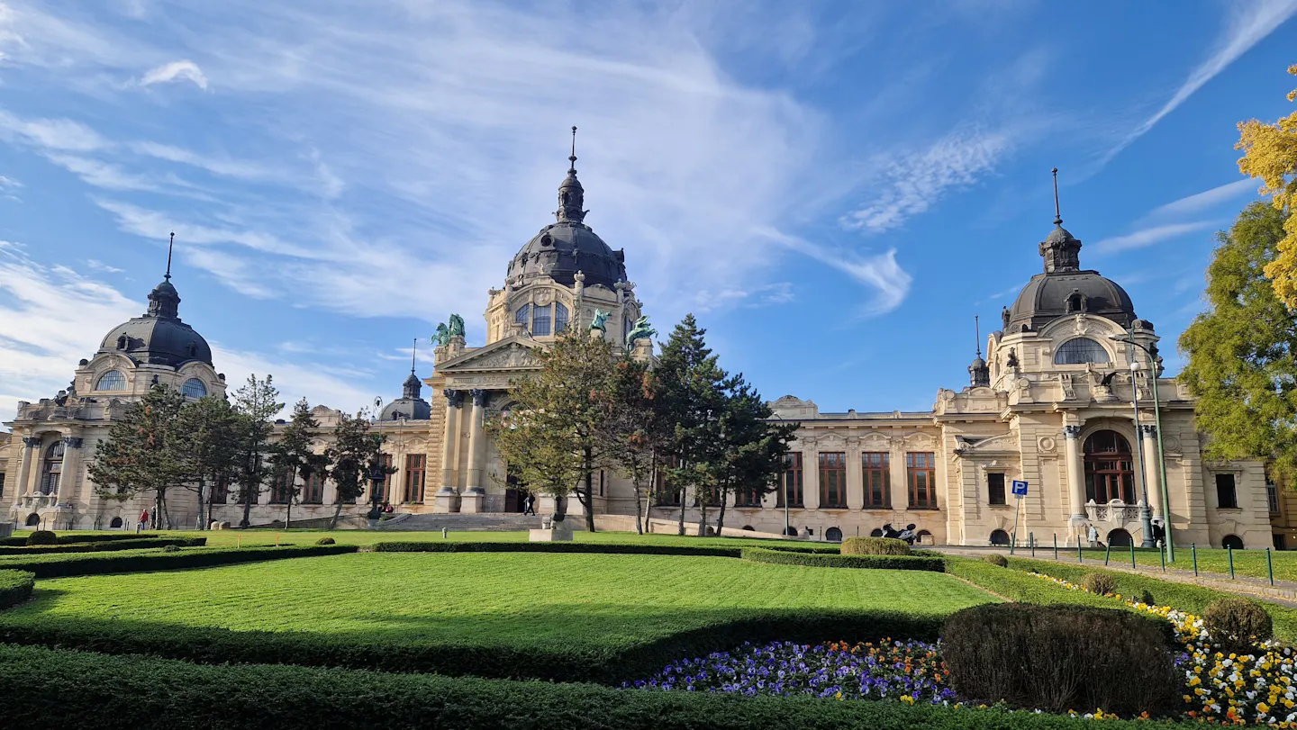 The domes and yellow facade of the Széchenyi thermal baths in Budapest above clipped lawns and flower beds