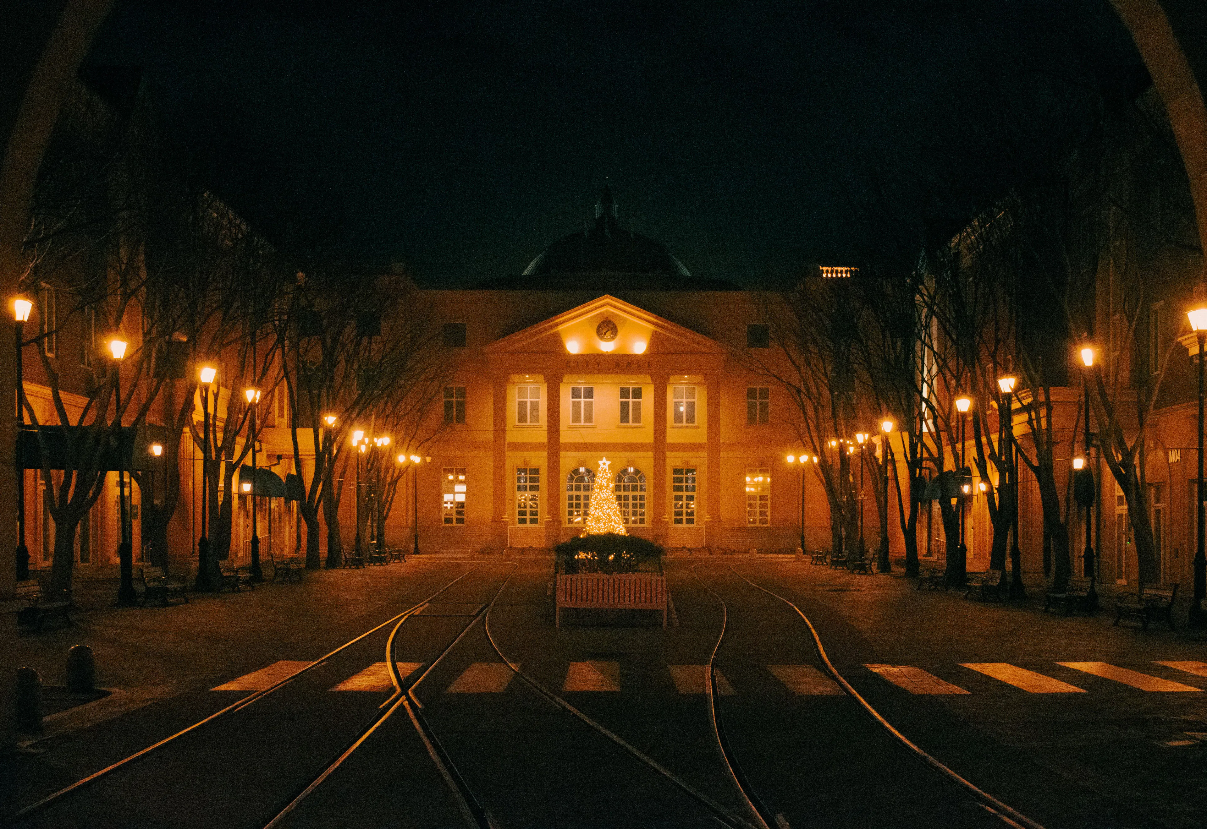 A lit Christmas tree in a quiet lamp-lit town square crossed by tram tracks at night
