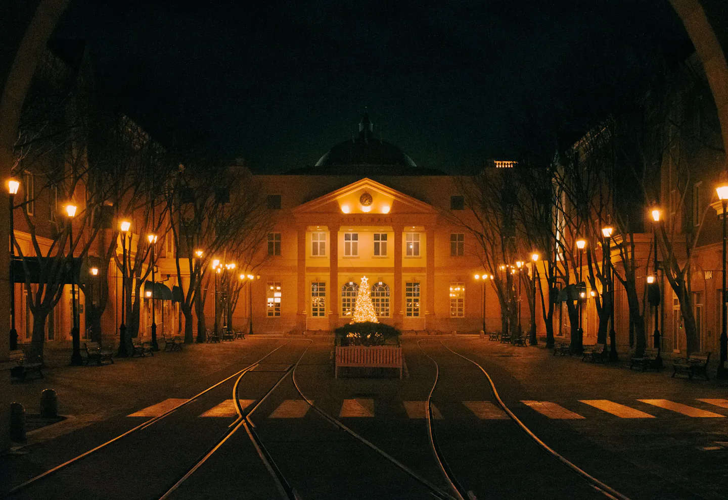 A lit Christmas tree in a quiet lamp-lit town square crossed by tram tracks at night
