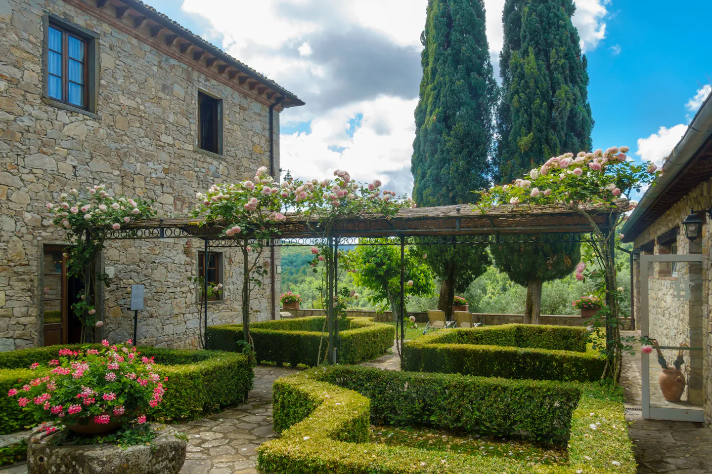 The walled garden of an Italian country villa: a wrought-iron pergola smothered in pale pink climbing roses over clipped box parterres, dry-stone walls and tall cypresses, under a blue sky with white cloud