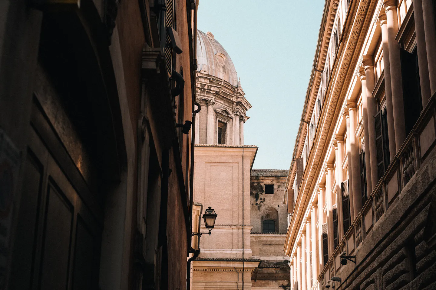 A baroque church dome and bell-stage rising above warm ochre and travertine palazzo facades that flank a narrow lane in Rome's historic centre