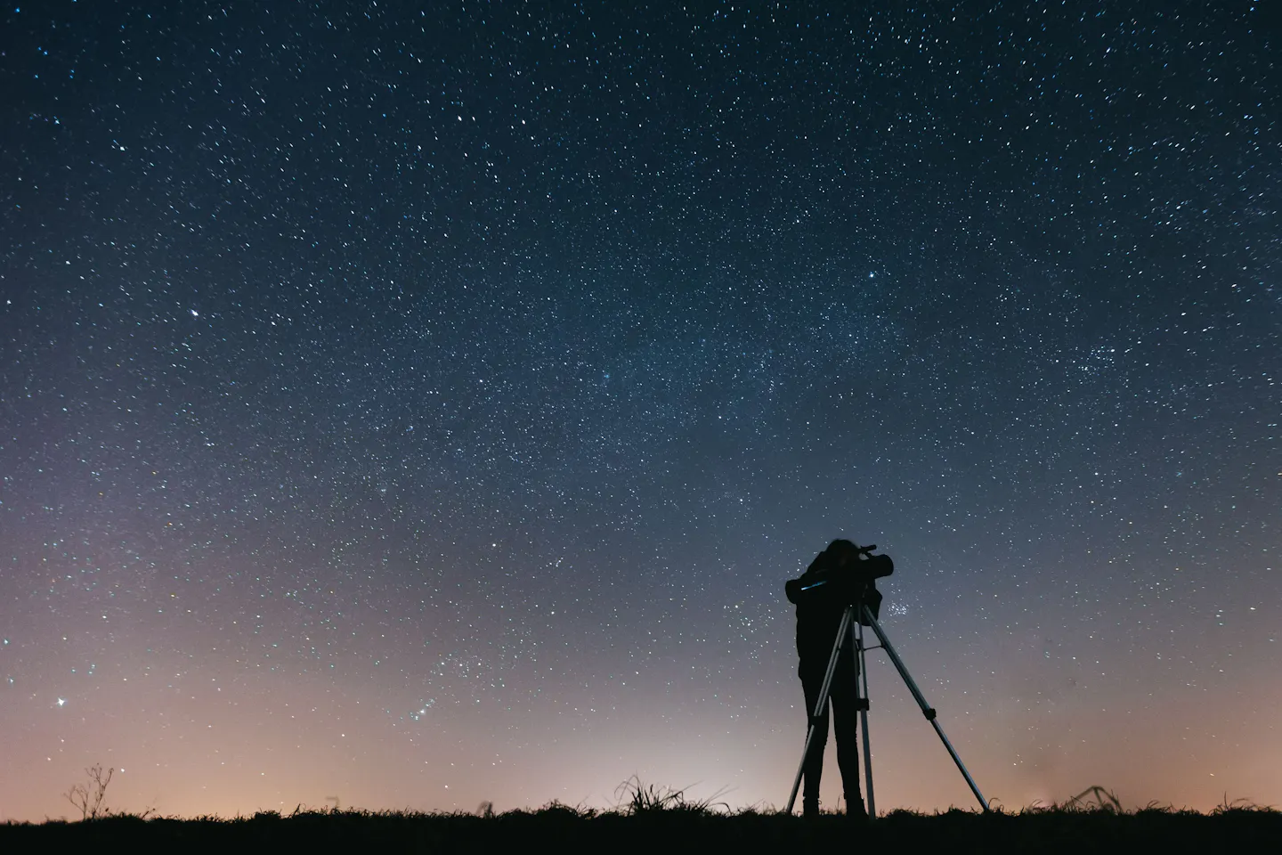 A stargazer silhouetted beside a telescope on open ground beneath a sky thick with stars, the last glow of dusk along the horizon