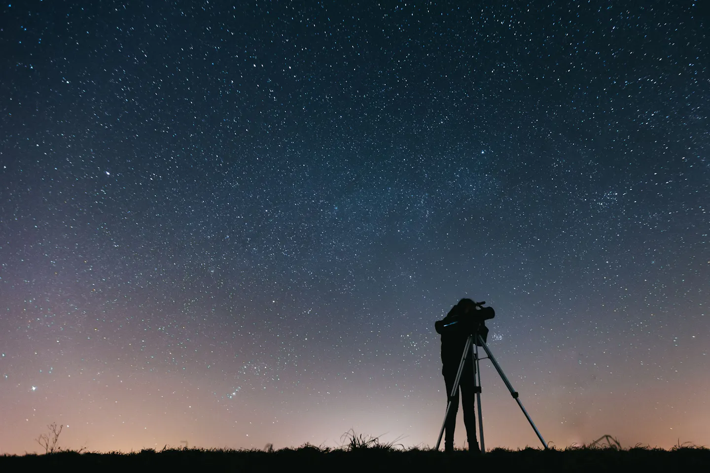 A stargazer silhouetted beside a telescope on open ground beneath a sky thick with stars, the last glow of dusk along the horizon