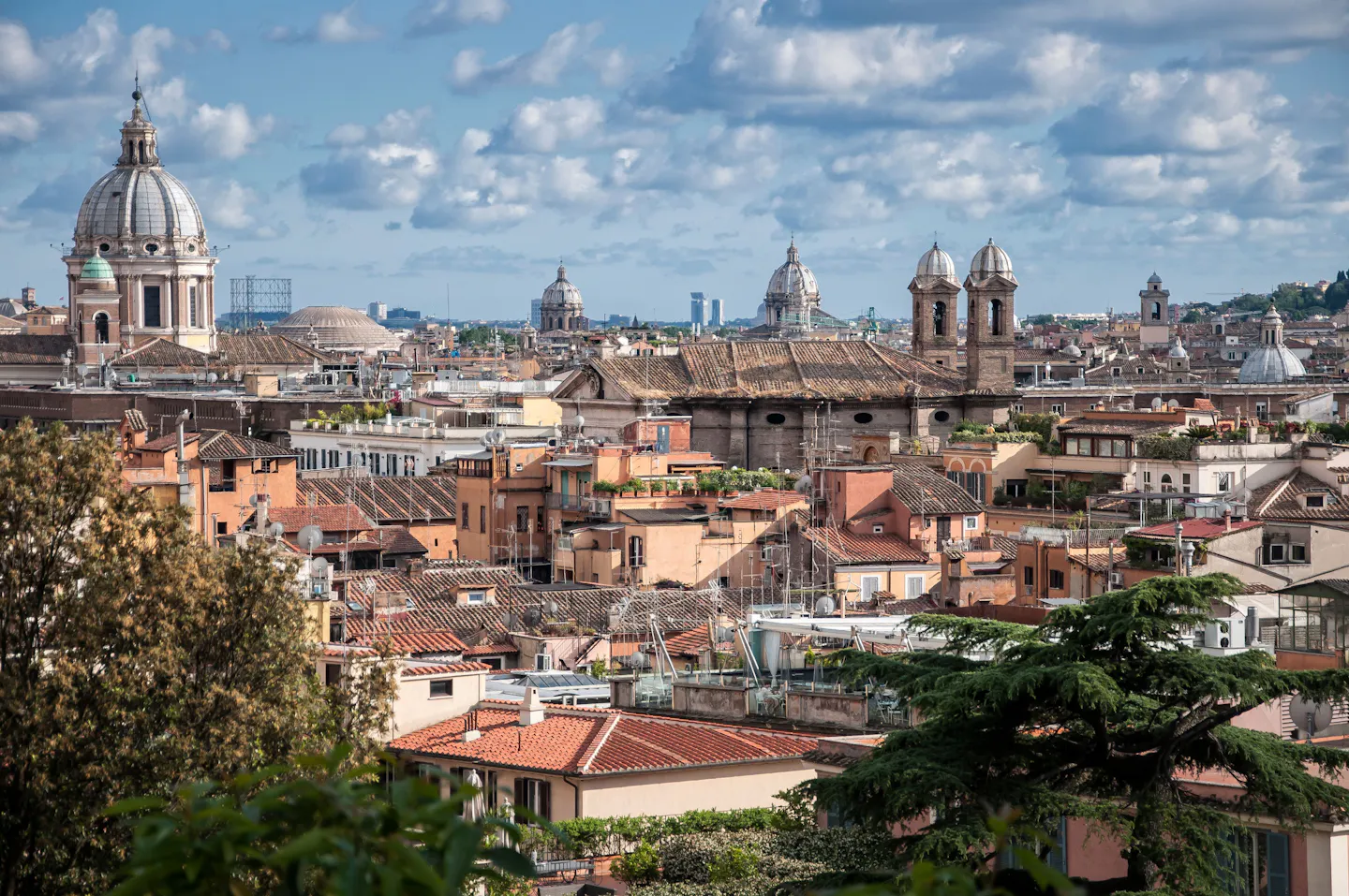 Terracotta rooftops, church domes and bell towers across central Rome under a blue sky with scattered cloud