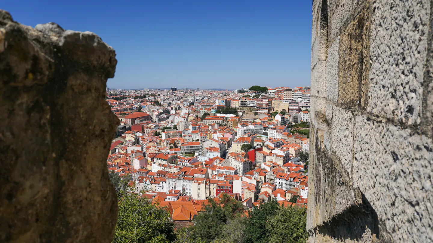 Lisbon's red-tiled rooftops spread out below, seen between two weathered stone merlons of the Sao Jorge Castle battlements under a deep blue sky