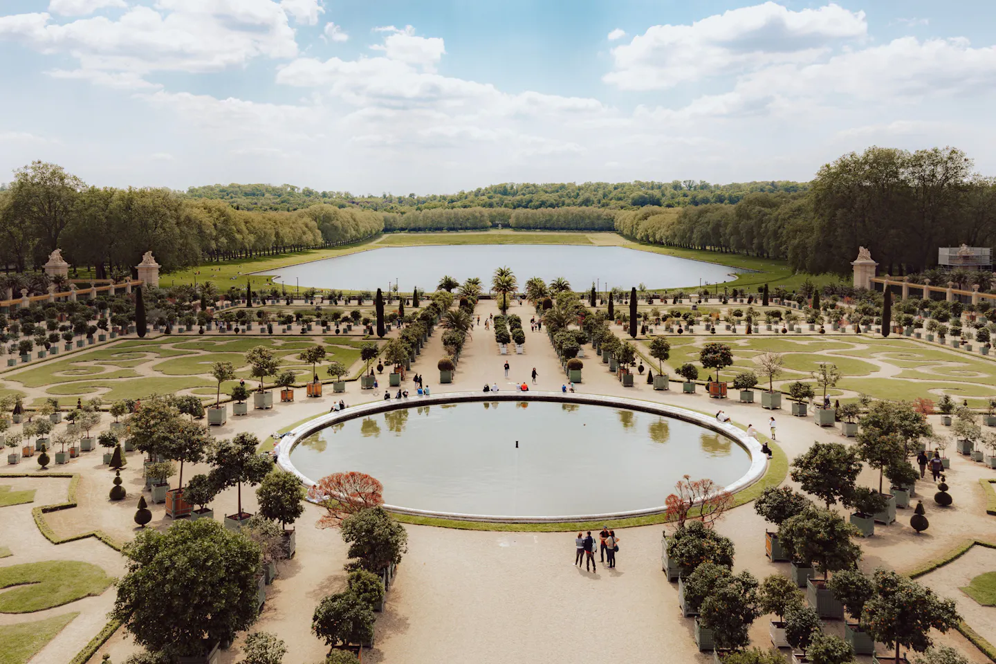Formal gardens at Versailles with clipped hedges, potted trees and a round pool, an ornamental lake beyond