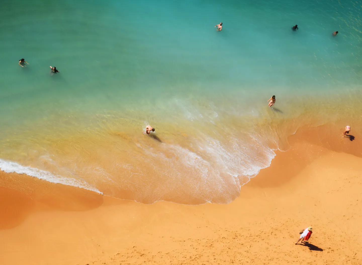 Swimmers in turquoise shallows along a curve of golden sand seen from above