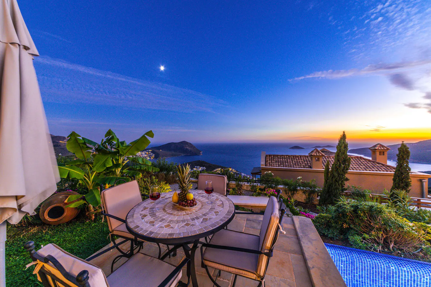 A terrace table set with wine and fruit above the Mediterranean as the sun sets behind the headlands