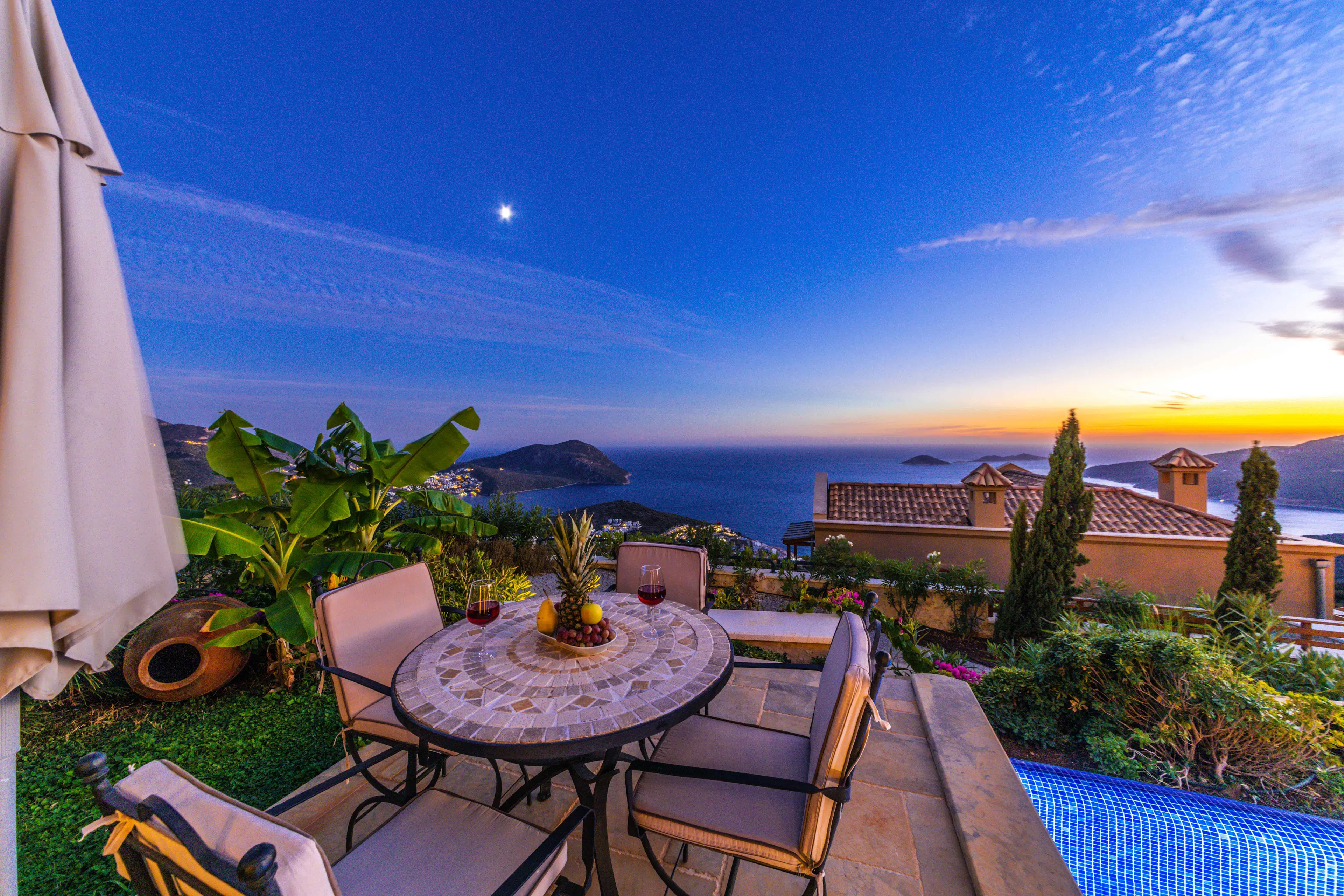 A terrace table set with wine and fruit above the Mediterranean as the sun sets behind the headlands