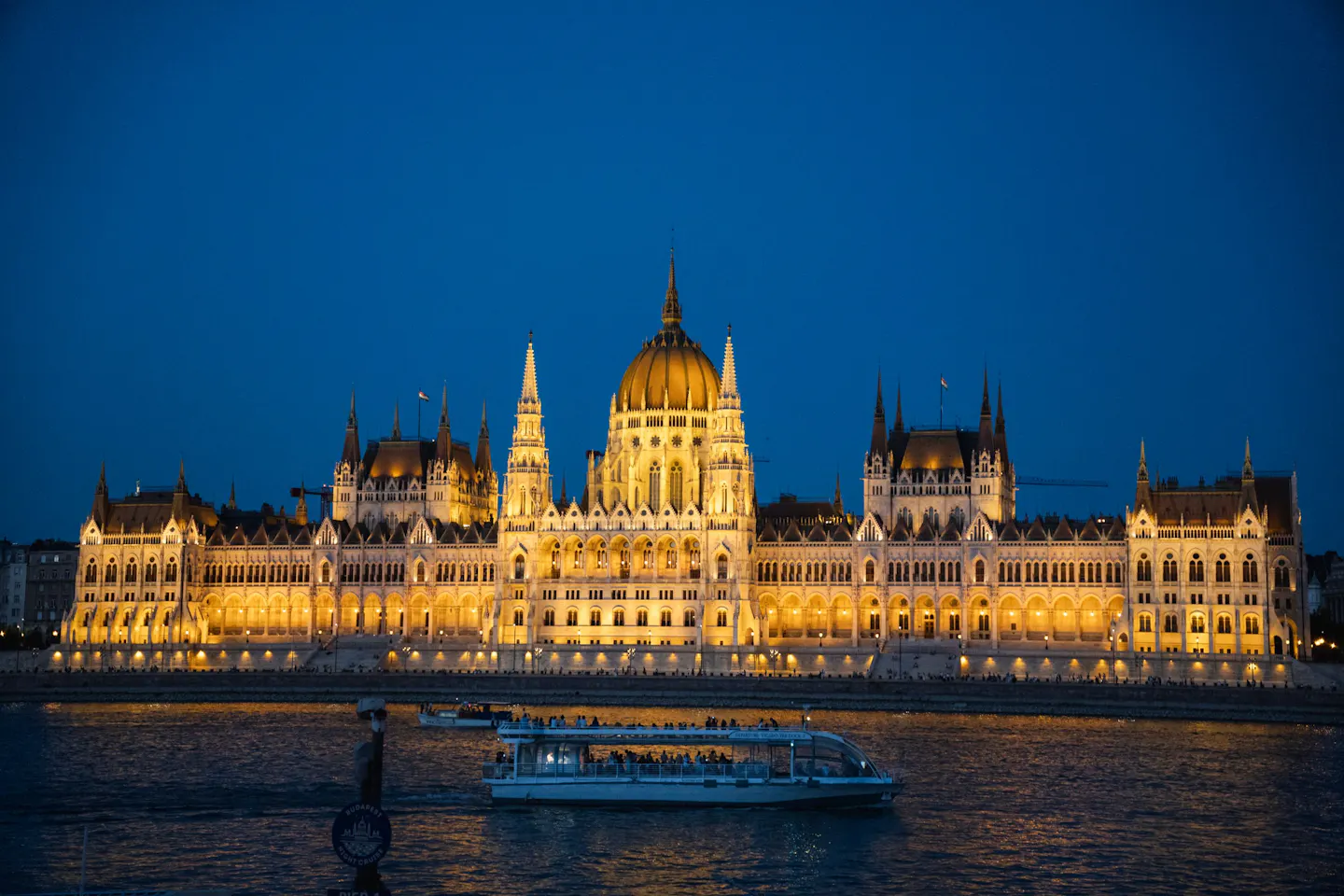 The Hungarian Parliament lit gold on the Danube at blue hour, with a boat passing in front