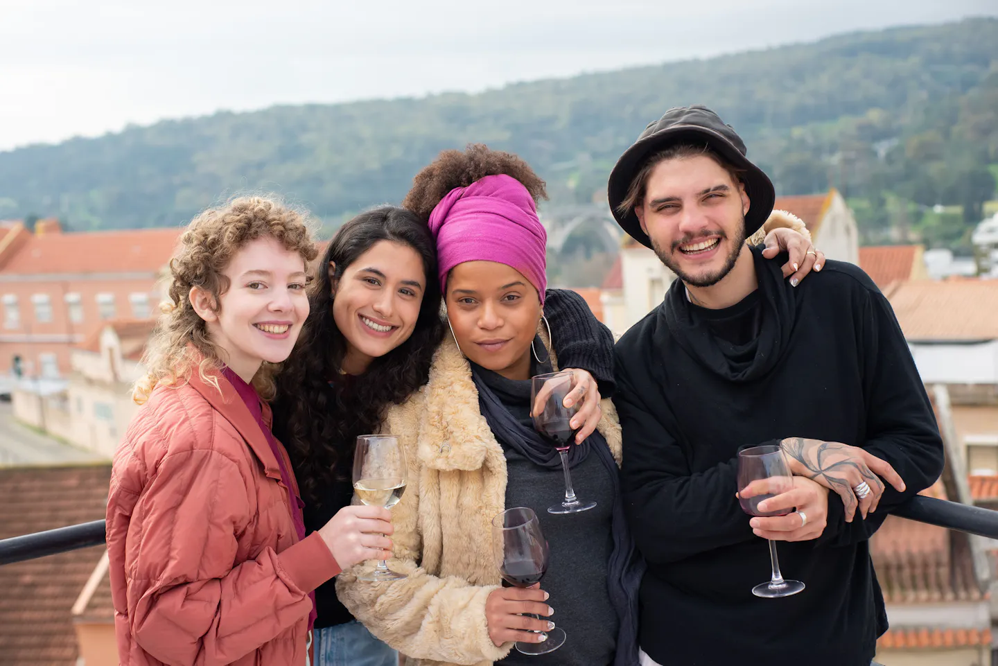 Four friends smiling together on a terrace overlooking a hilly, red-roofed city