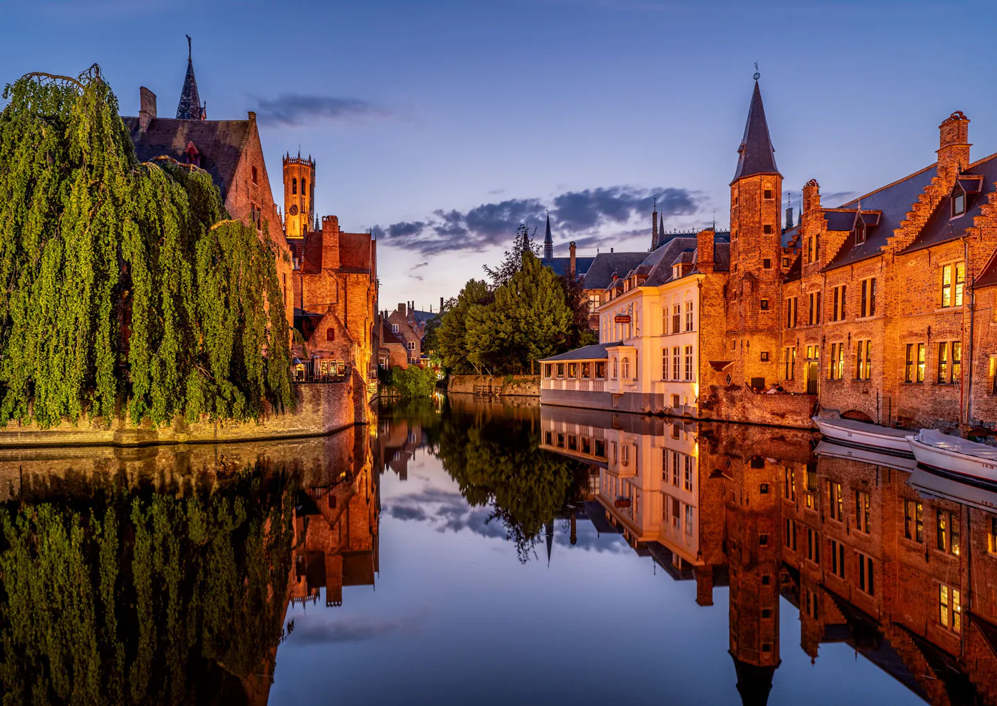 Lamplit brick houses and a weeping willow mirrored in a still Bruges canal at twilight