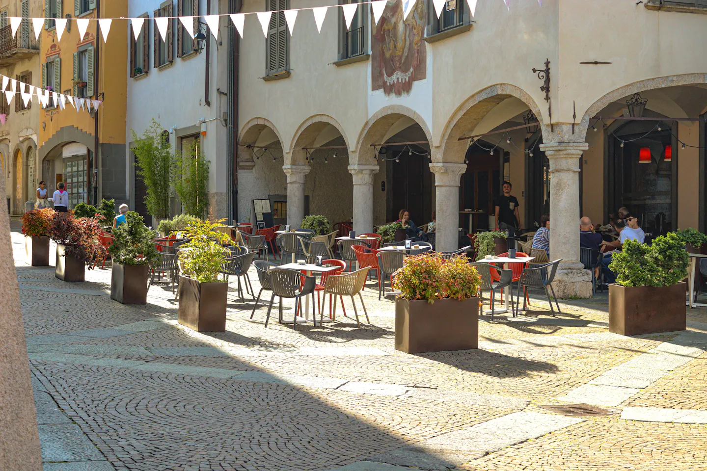 Cafe tables and potted shrubs on a sunlit cobbled piazza beneath stone arcades in Italy