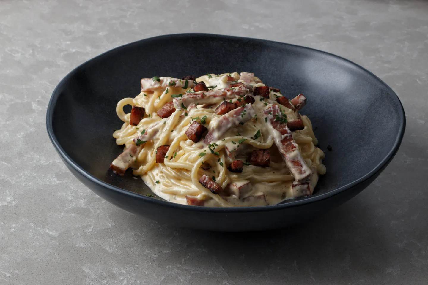 A bowl of spaghetti carbonara with crisp cubes of guanciale and chopped herbs in a dark ceramic bowl
