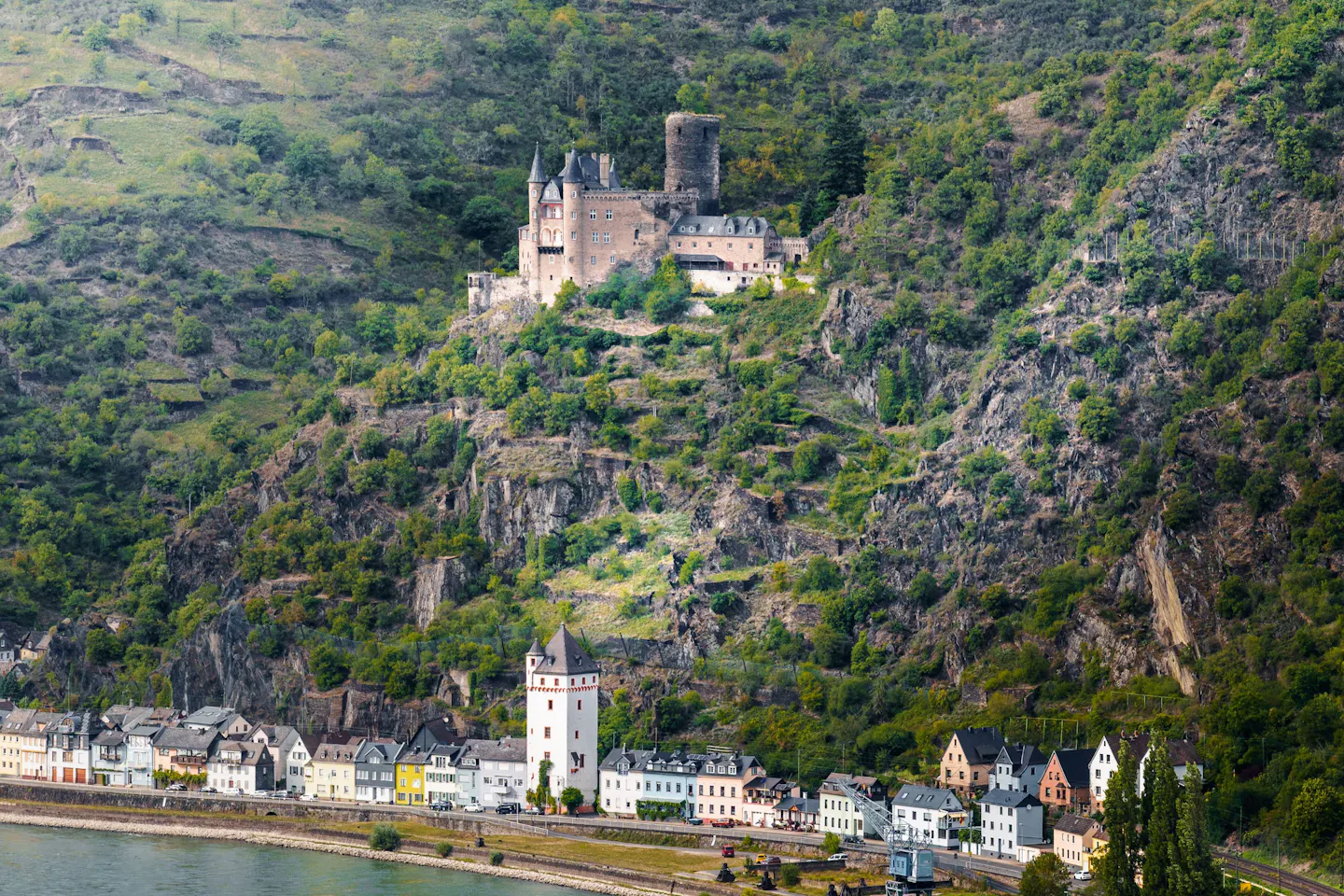 A hilltop castle above a riverside village and the railway line on the steep green banks of the Rhine