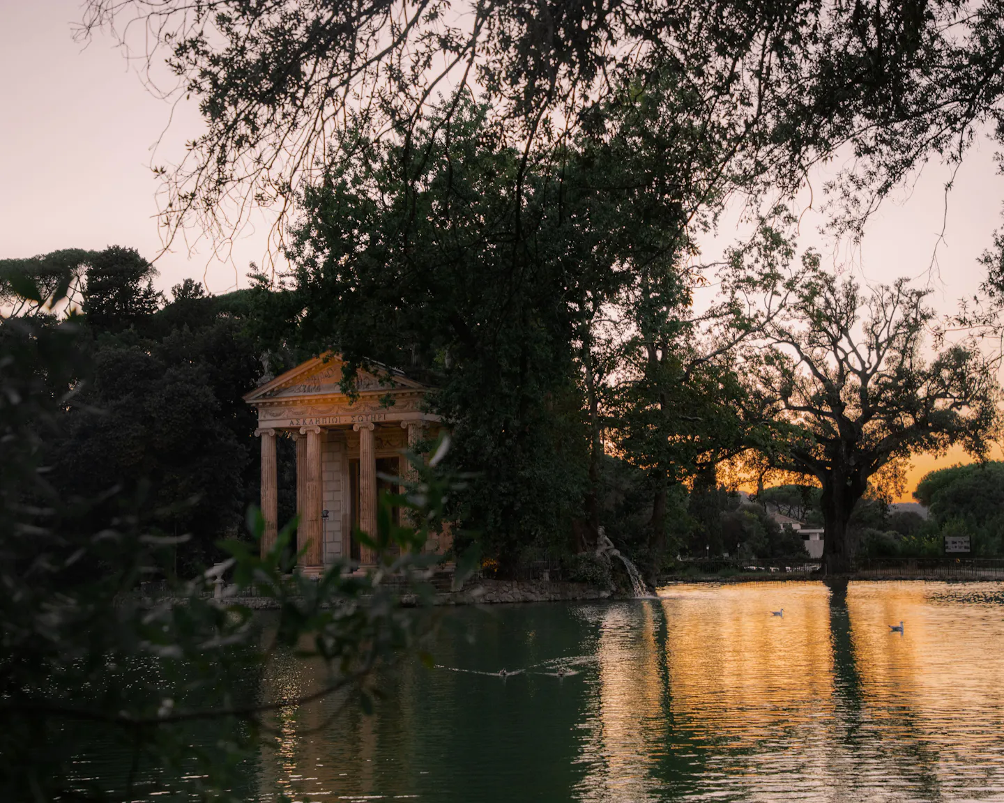 The columned Temple of Aesculapius beside the lake in Villa Borghese at sunset, framed by dark foliage with golden light on the water