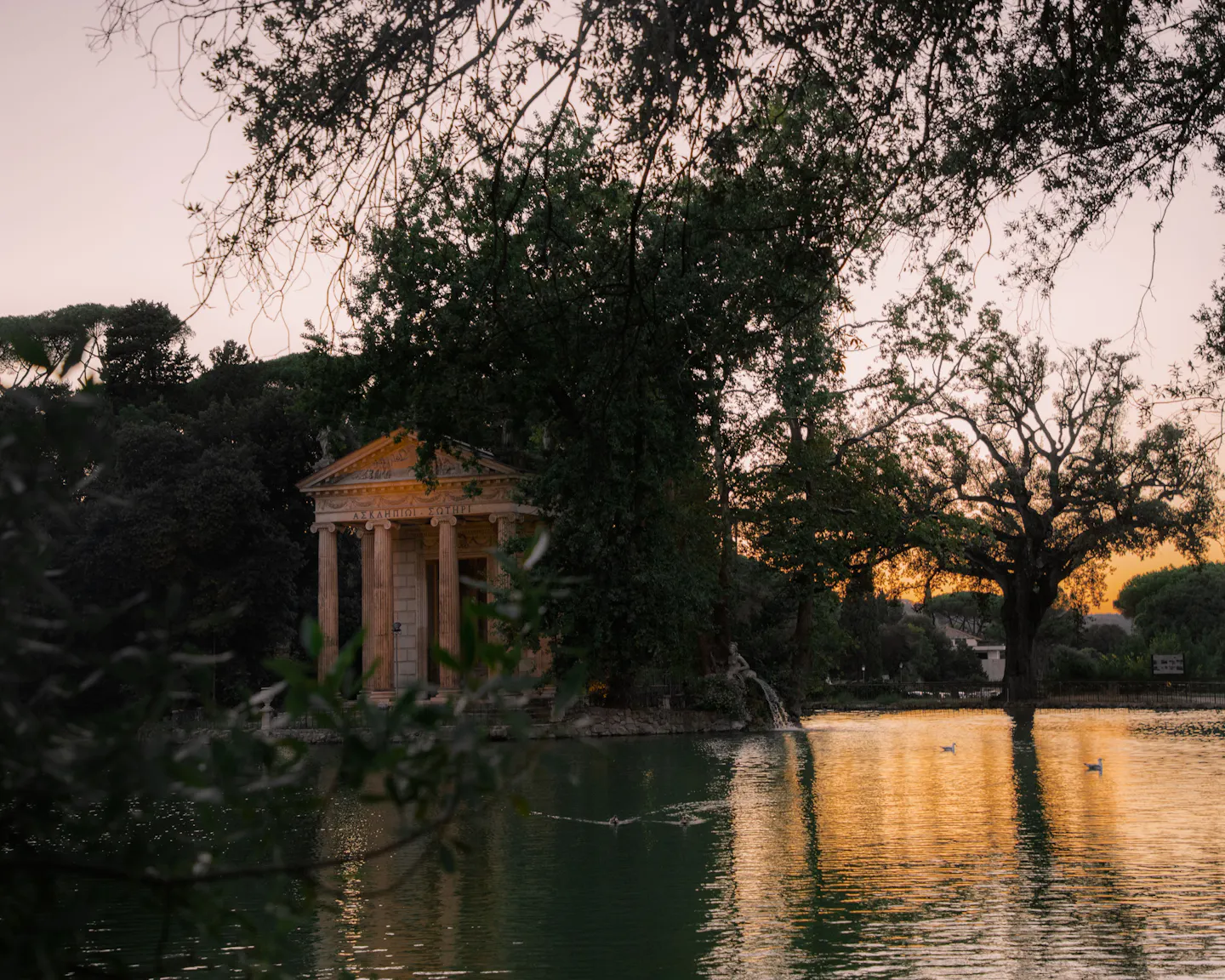 The columned Temple of Aesculapius beside the lake in Villa Borghese at sunset, framed by dark foliage with golden light on the water