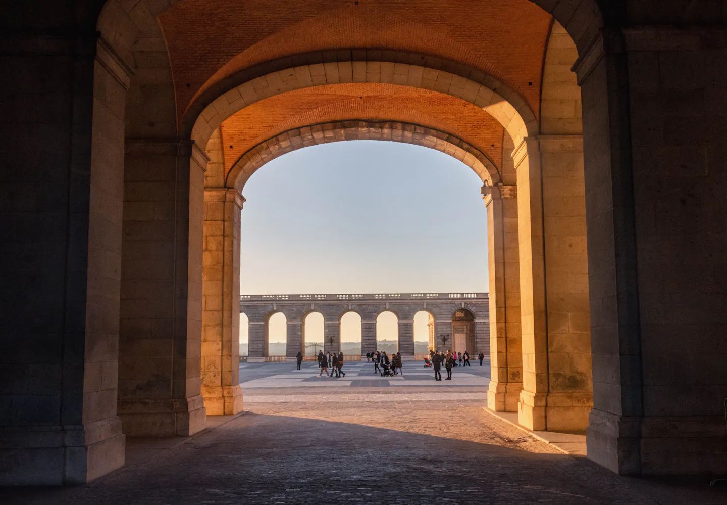 A sunlit palace courtyard in Madrid framed by a stone archway, with people crossing in front of the colonnade
