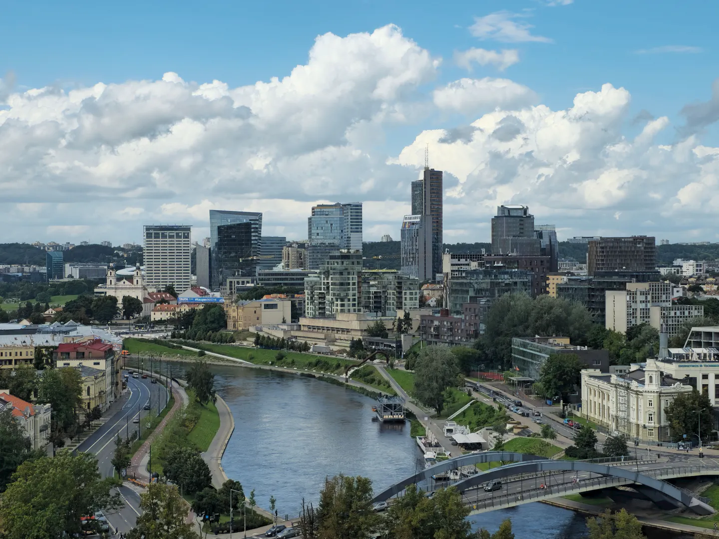 Glass office towers of the modern Vilnius business district rising across the Neris River from green banks